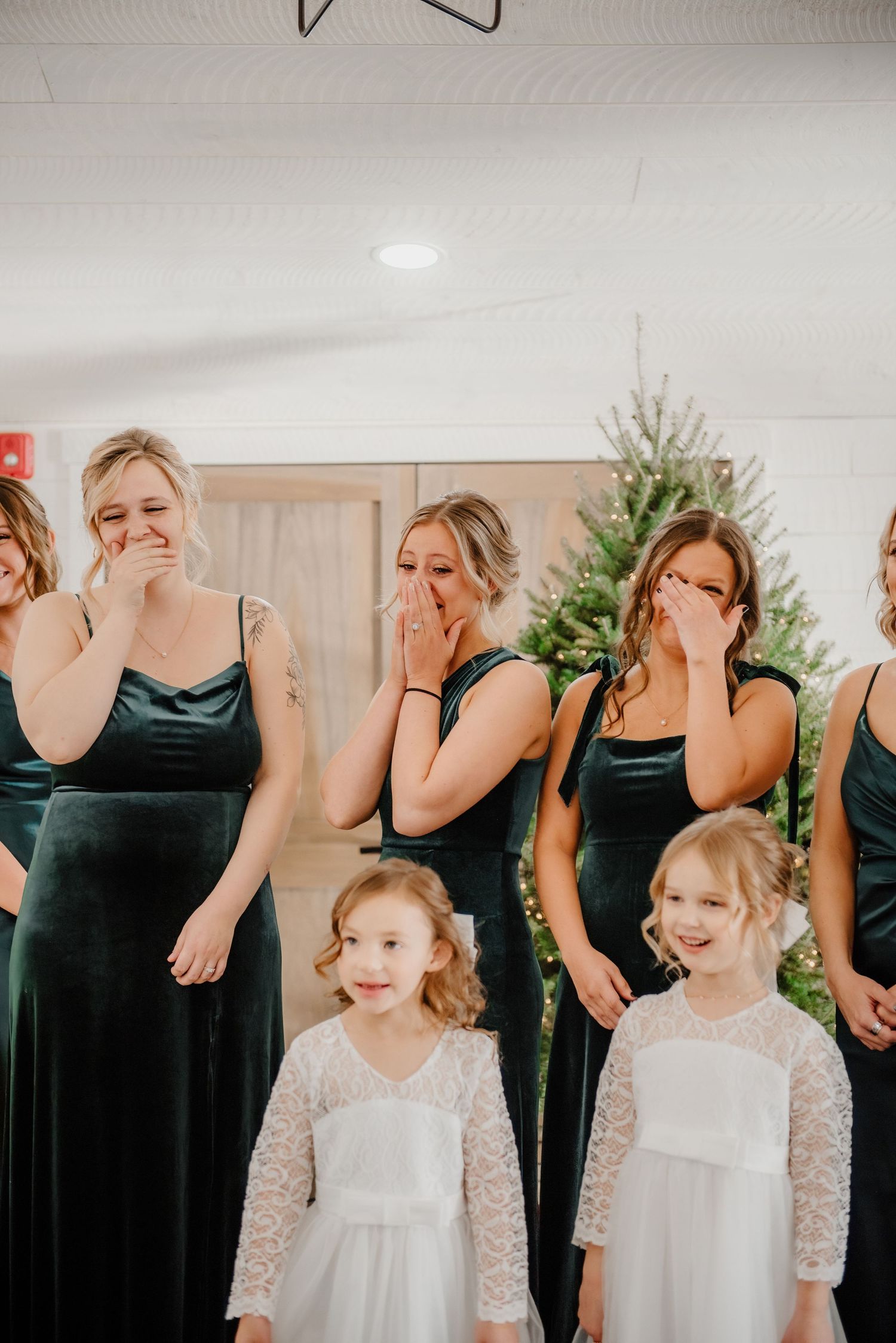 Bridesmaids in green velvet dresses react emotionally with flower girls in white lace dresses at a Christmas wedding.