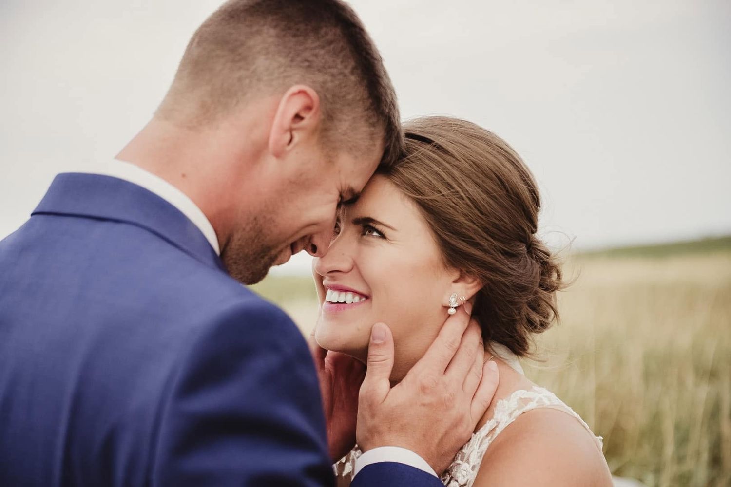 A smiling bride and groom share a tender forehead-to-forehead moment in a golden field on their wedding day.