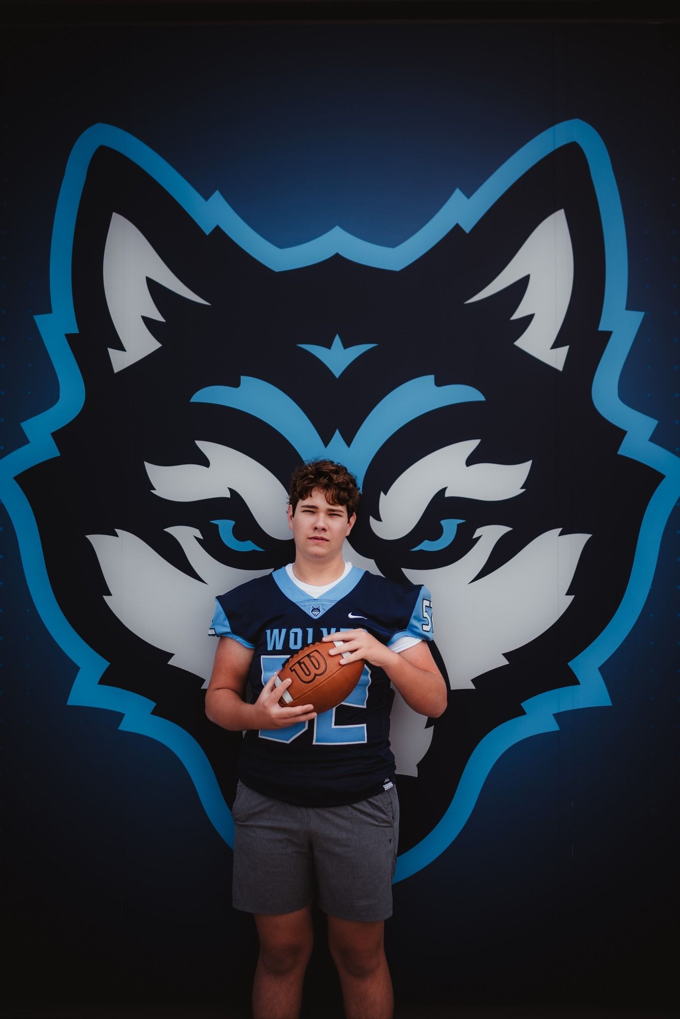 Young football player in Wolves jersey holding a football, posing in front of a blue wolf mascot logo.