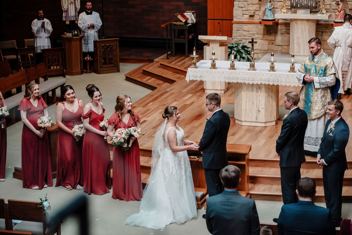Bride and groom exchange vows at church altar surrounded by bridesmaids in red dresses during elegant wedding ceremony.