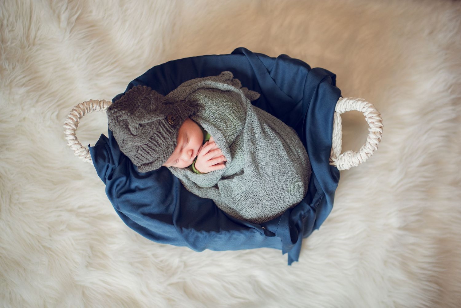 Newborn baby sleeping in a basket wrapped in a gray knit swaddle and matching bear hat on a white fur rug.