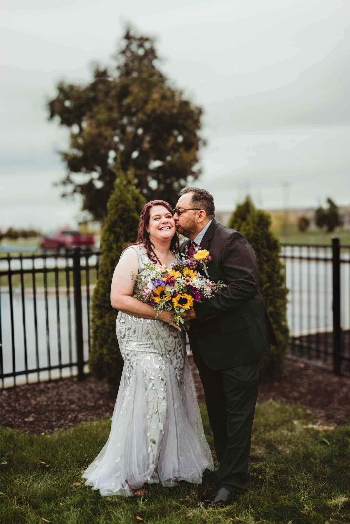 A bride in a silver gown holds a sunflower bouquet while her groom kisses her cheek outdoors at their wedding.