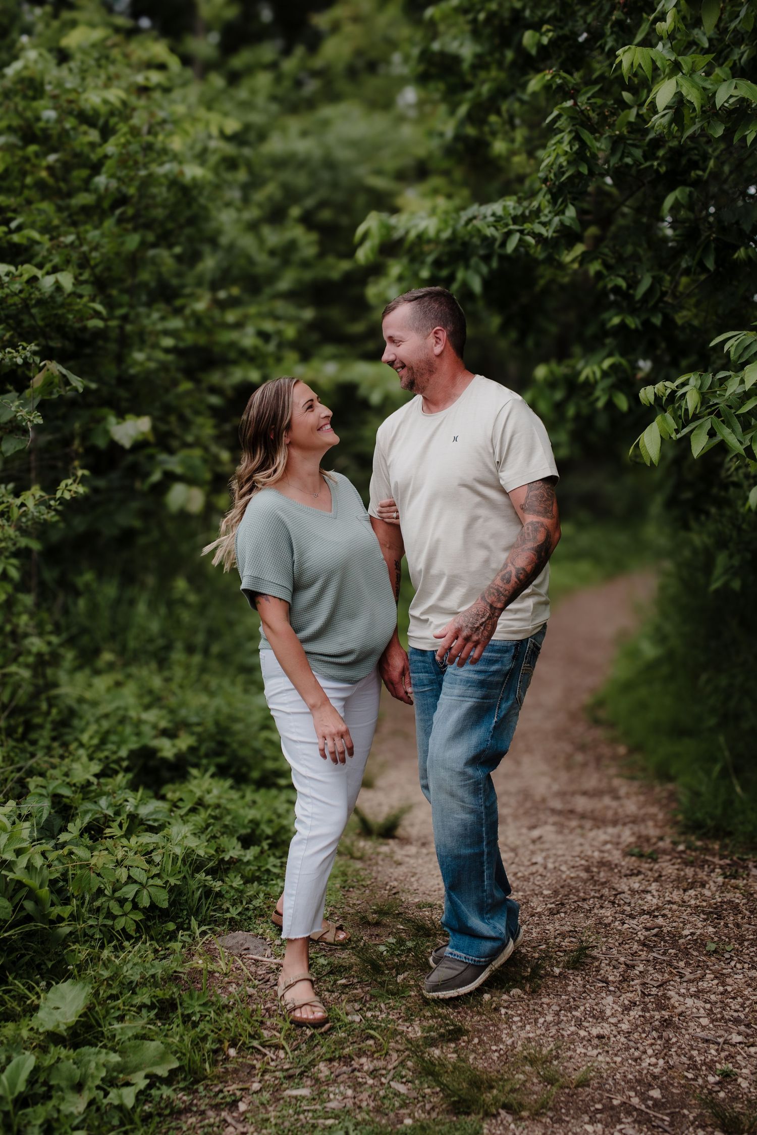 A smiling couple walks hand-in-hand on a lush green nature trail, wearing casual outfits of sage green and cream tops.