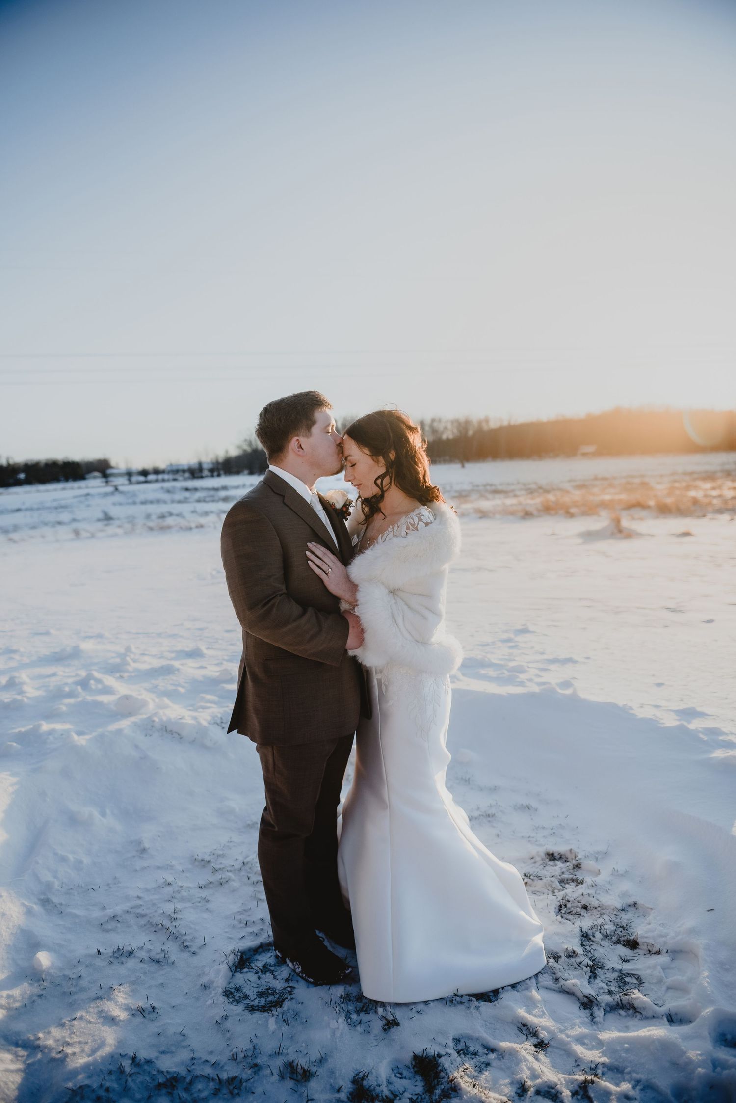 Bride and groom share a kiss in a snowy winter field at golden hour, bride in white gown, groom in brown suit.