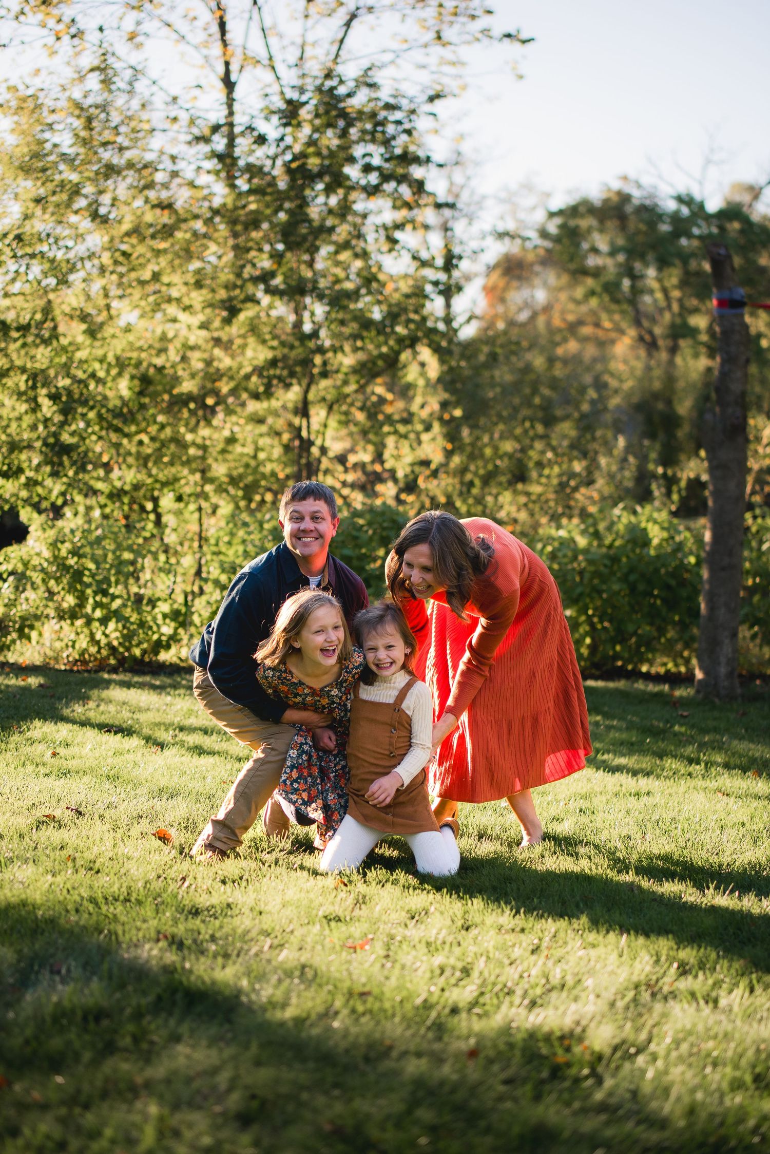 Family of four laughing together outdoors on green grass surrounded by autumn trees in warm fall sunlight.