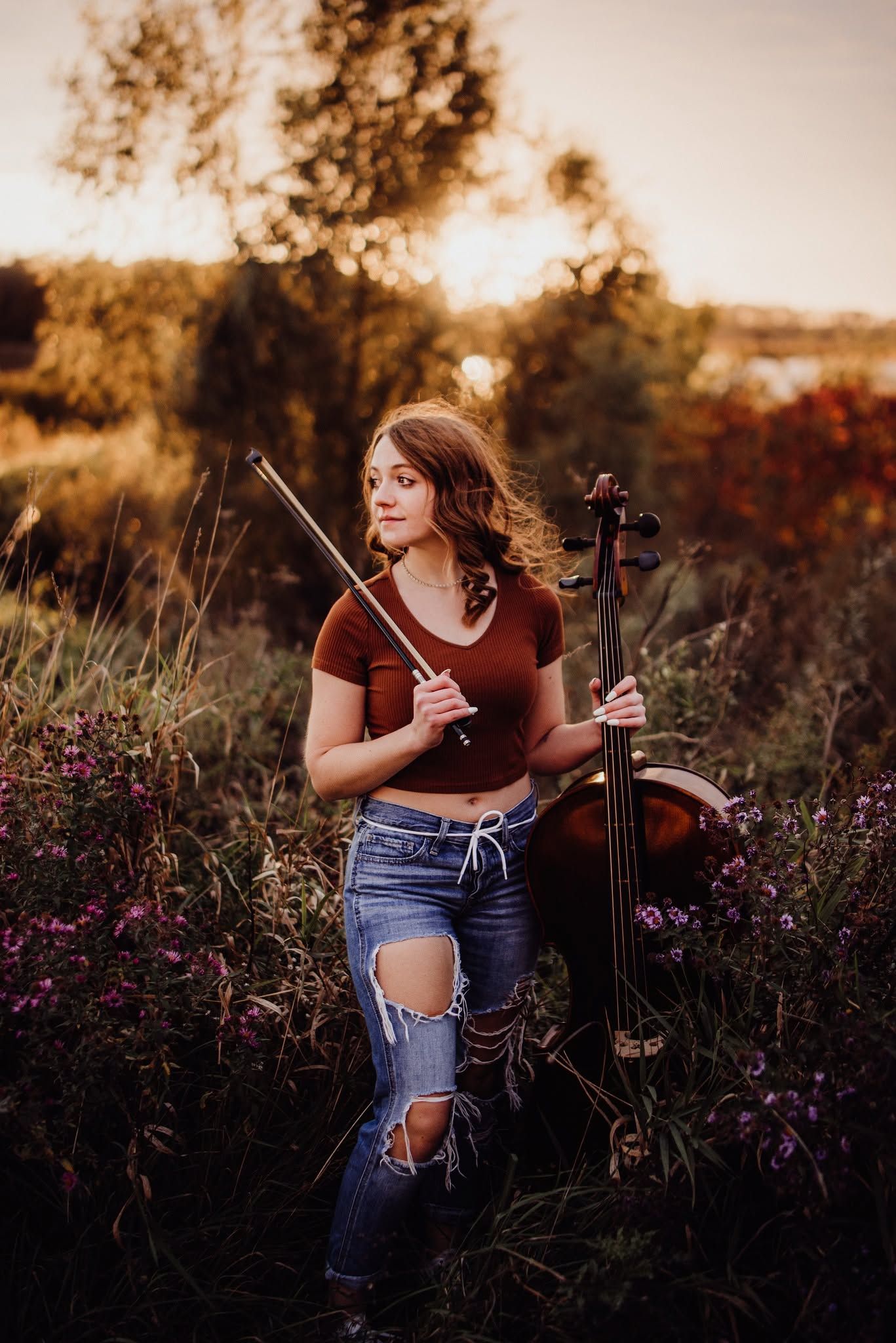 Young woman holding a cello and bow in a wildflower field at golden hour sunset.
