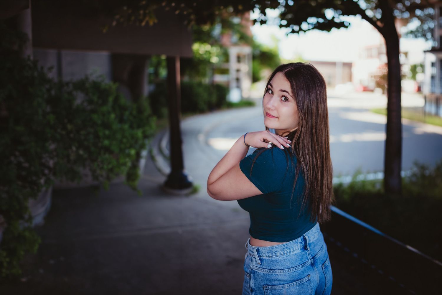 Young woman in blue crop top and jeans poses outdoors on a tree-lined walkway, glancing back over her shoulder.