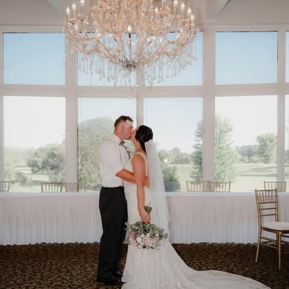 A bride and groom share a kiss under an elegant crystal chandelier in a bright wedding venue with large windows.