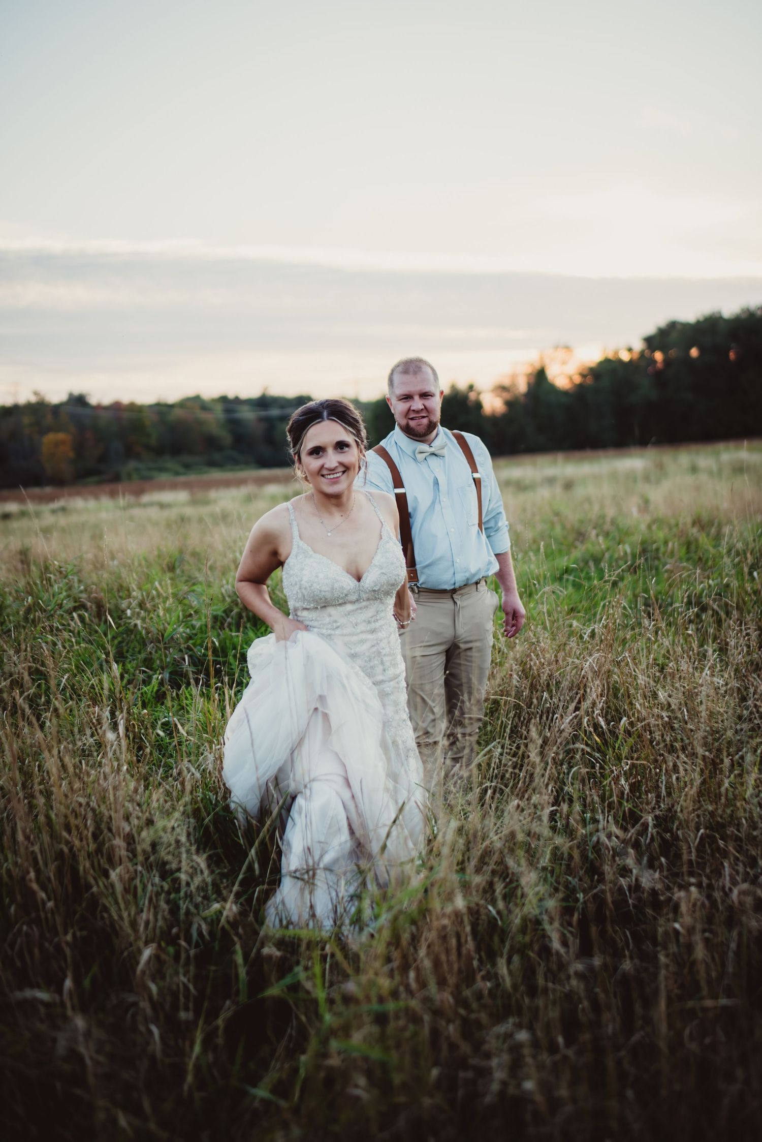 Bride in white lace gown and groom in suspenders walk through golden field at sunset.