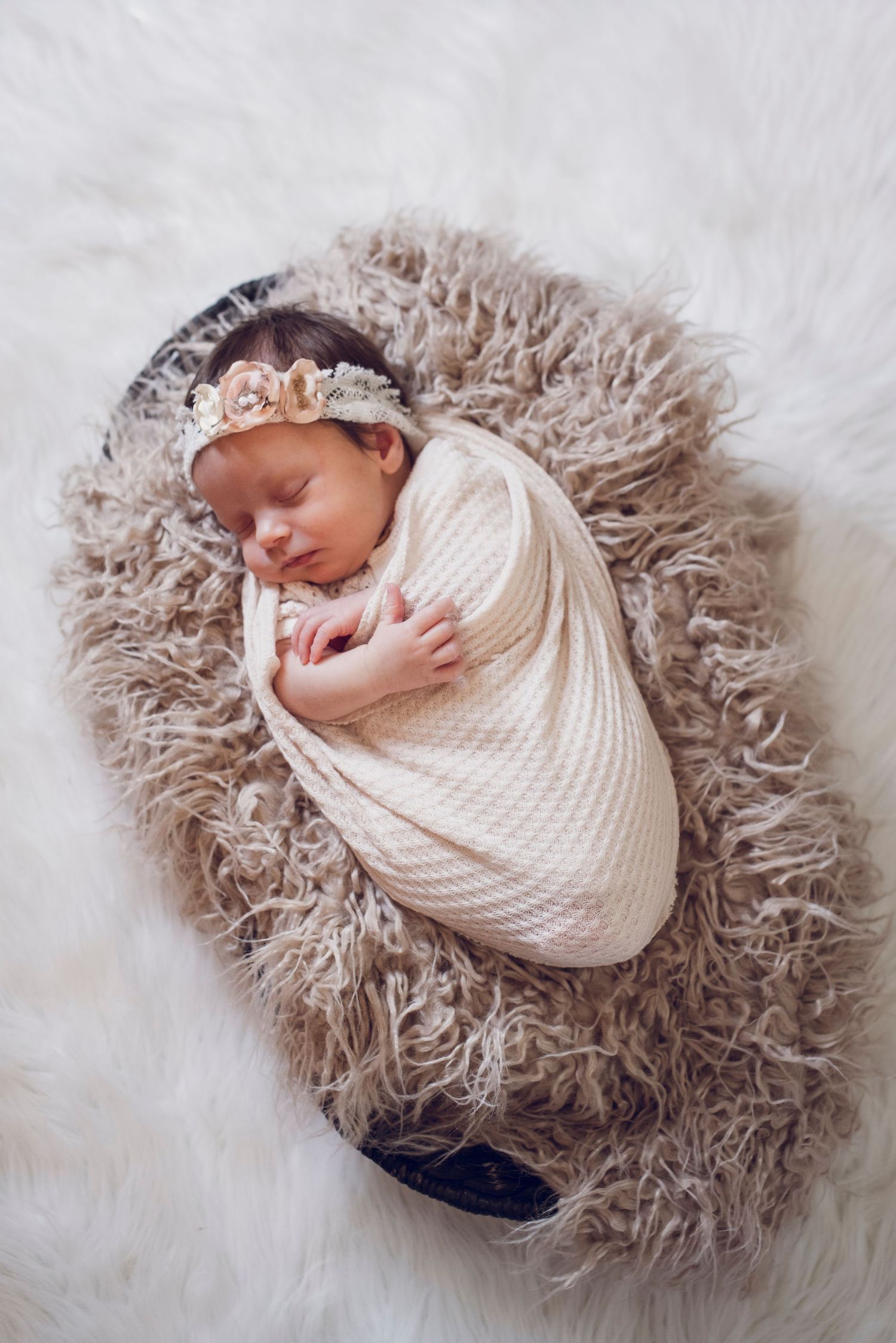 Newborn baby swaddled in cream wrap with floral headband sleeping in fluffy brown basket on white fur.