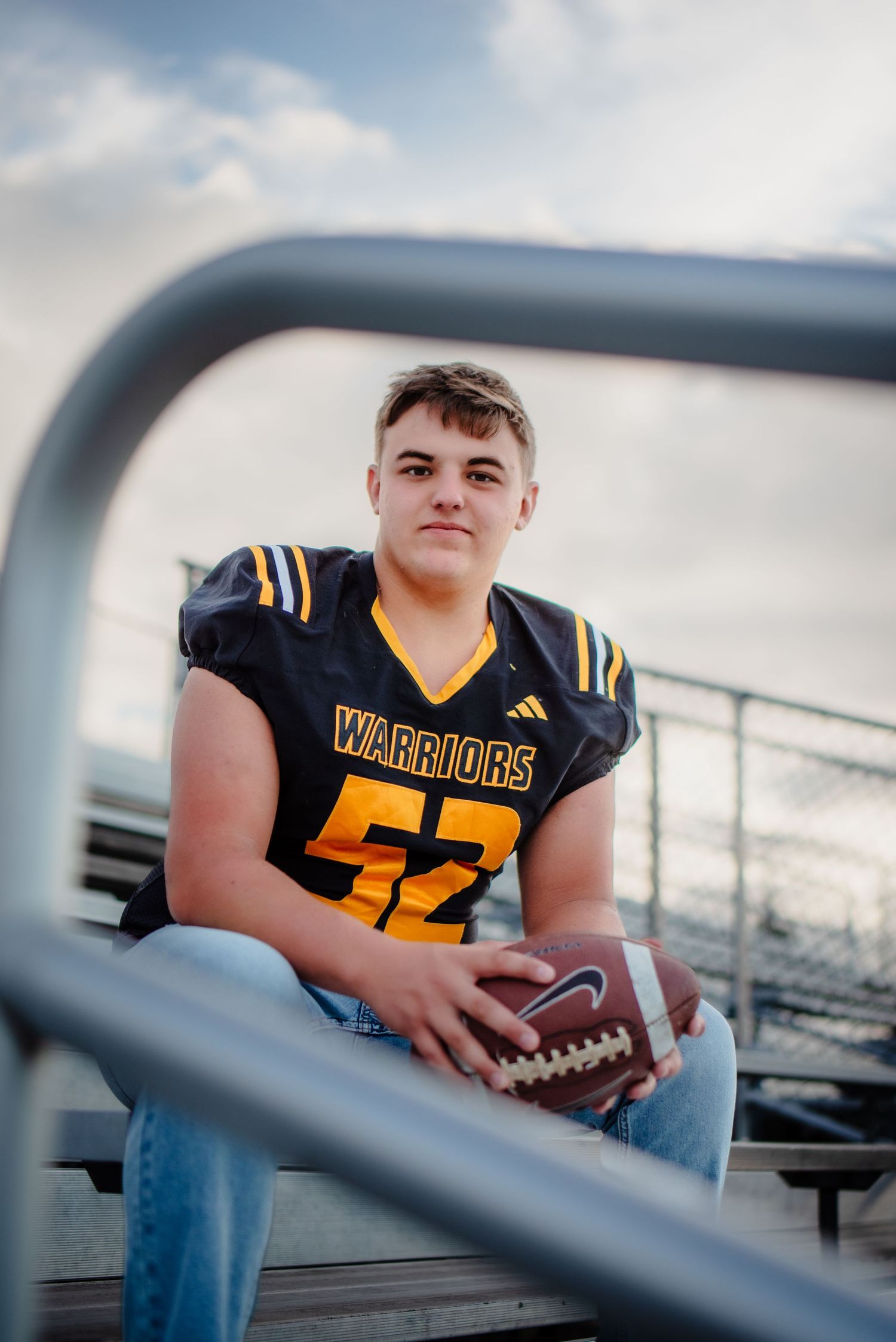 Young football player in Warriors jersey #52 sits on bleachers holding a football during senior portrait session.