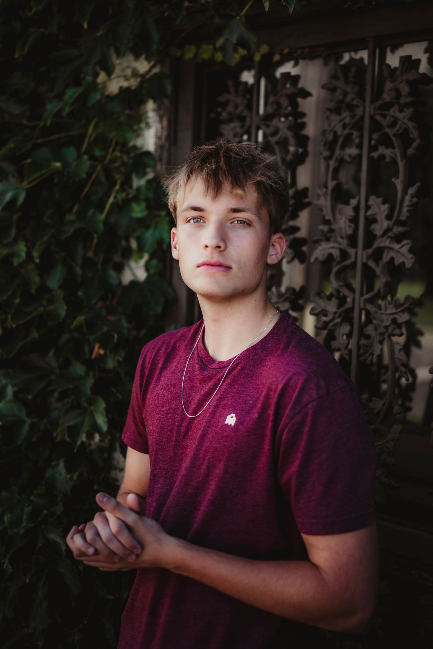 Young man in burgundy t-shirt with chain necklace poses outdoors near ivy-covered decorative iron gate.