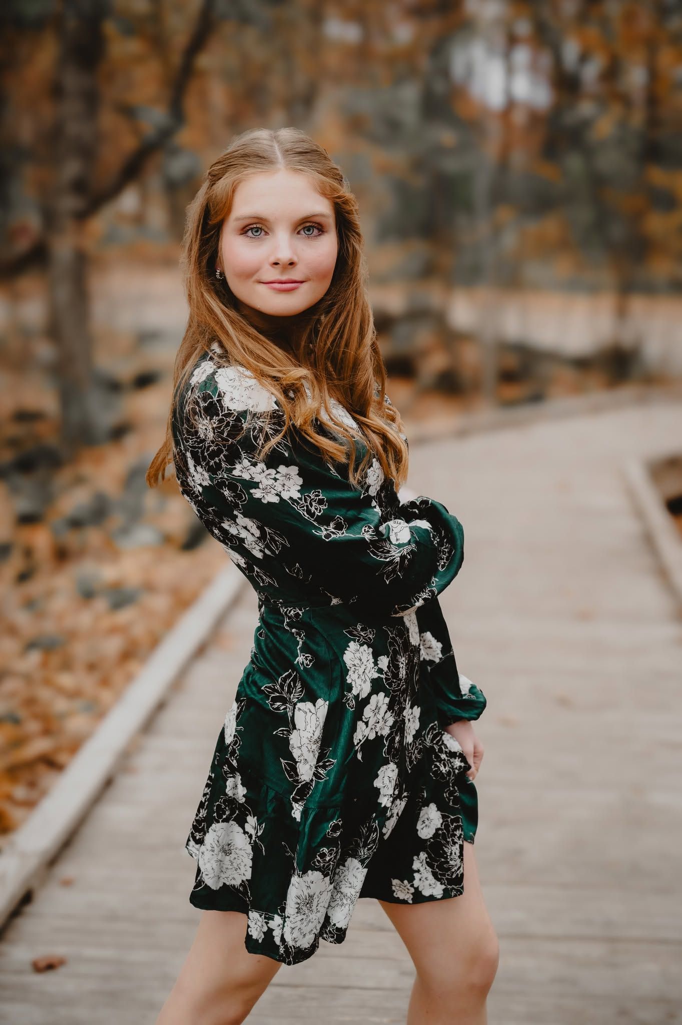 Young woman in green floral dress poses on wooden path surrounded by autumn foliage.