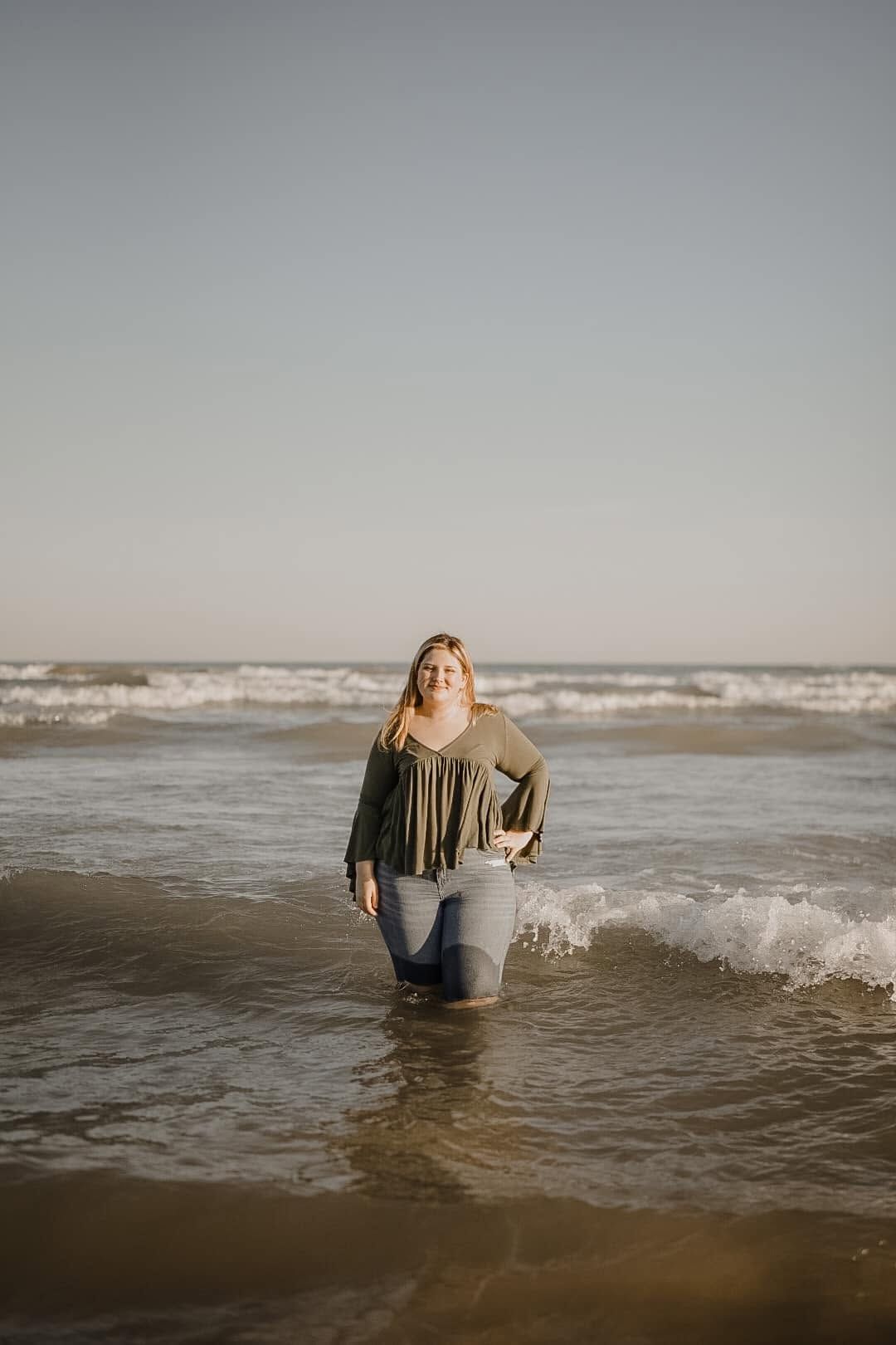 Woman in green top and jeans wading in ocean waves on a sandy beach at golden hour.