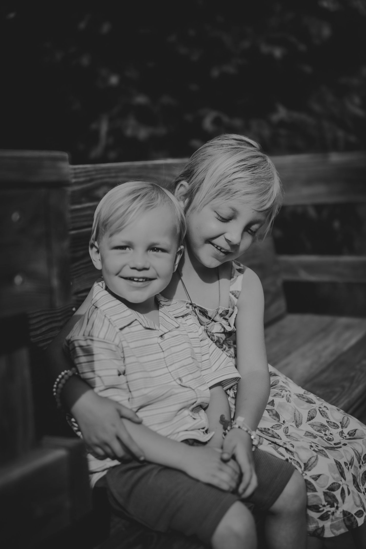 Black and white photo of two young blonde siblings hugging and smiling on a wooden bench outdoors.