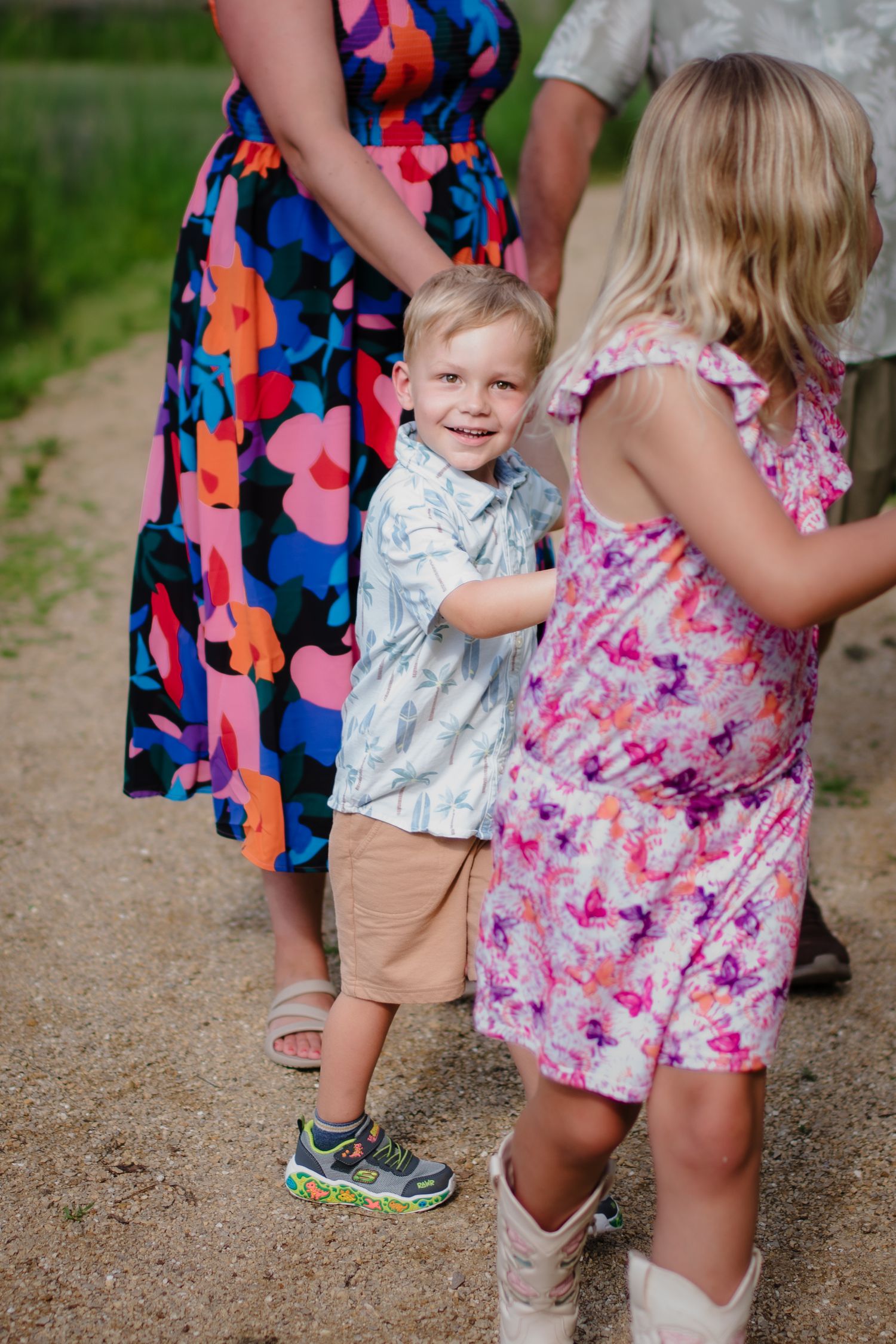 Young boy smiling on a gravel path with family, wearing a floral shirt and khaki shorts during an outdoor photoshoot.