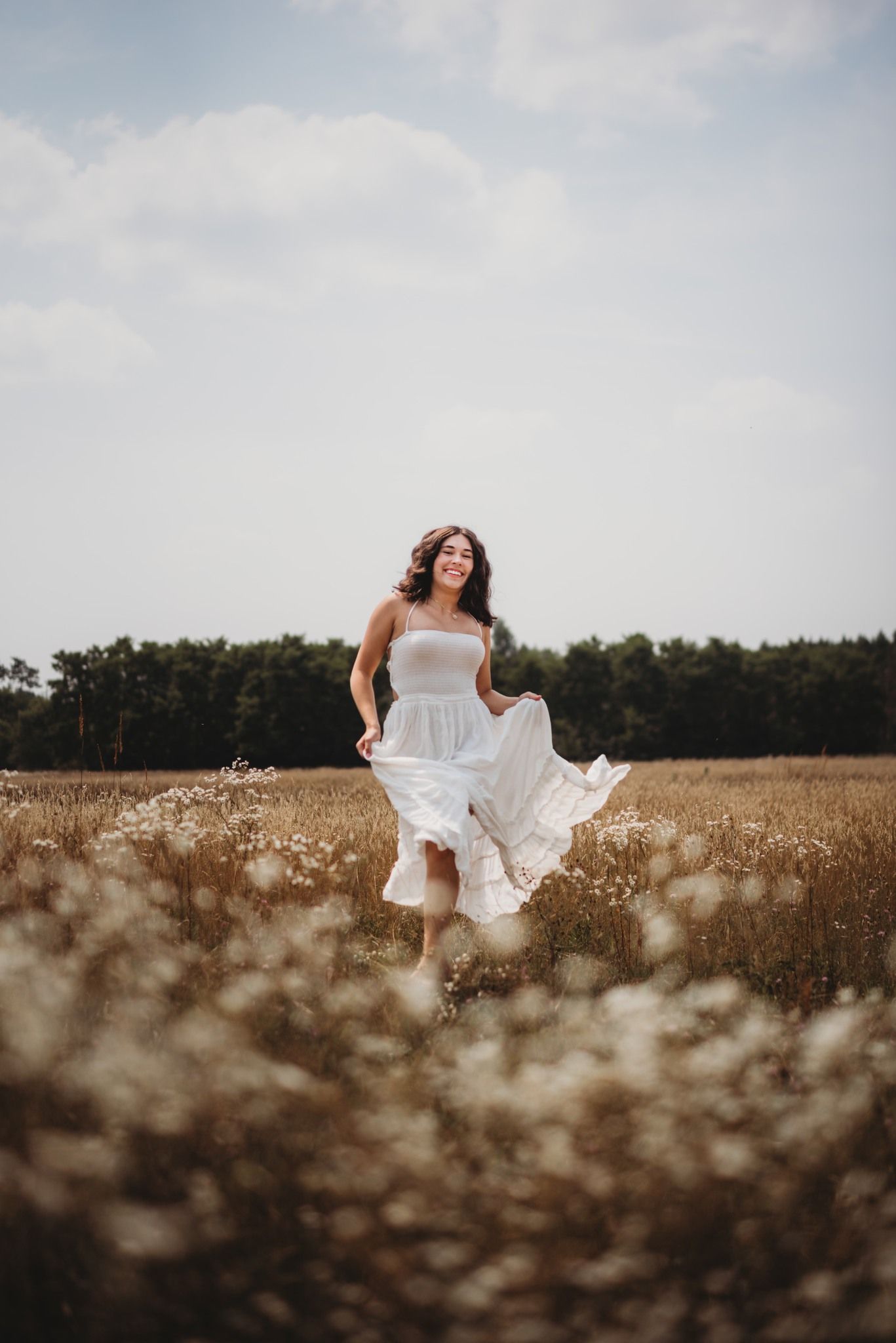 Woman in white dress running through a golden wildflower field with trees in background on cloudy day.