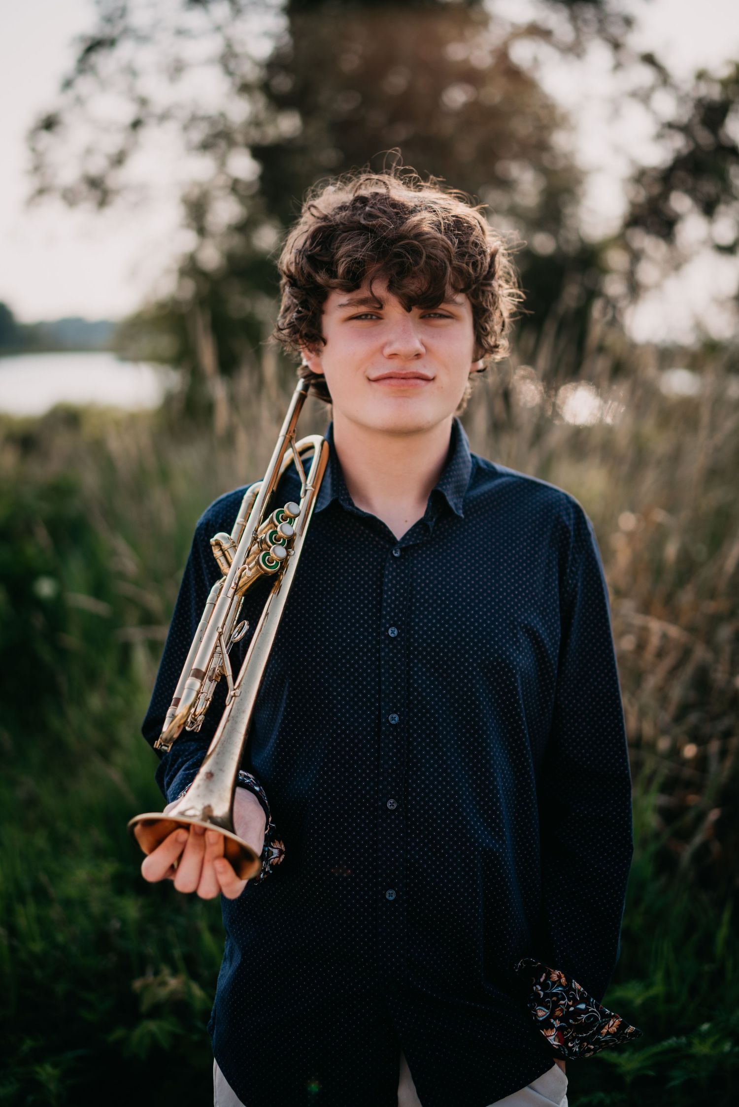 Young musician holding a trumpet in a field, wearing a dark blue shirt, with natural greenery in the background.