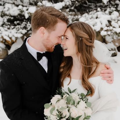 A bride and groom share a romantic moment in a snowy winter setting, with the bride holding a white rose bouquet.