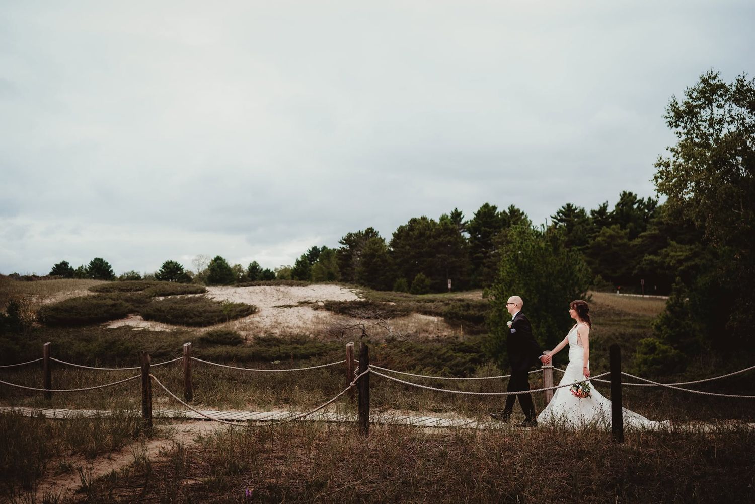 Bride and groom walk hand-in-hand along a rustic rope fence with sandy dunes and pine trees in the background.