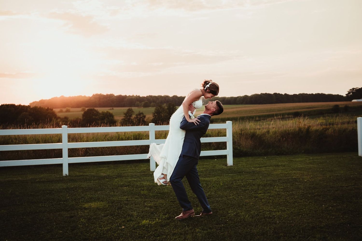 Groom lifts bride in white dress at sunset by white fence on green lawn during outdoor wedding celebration.