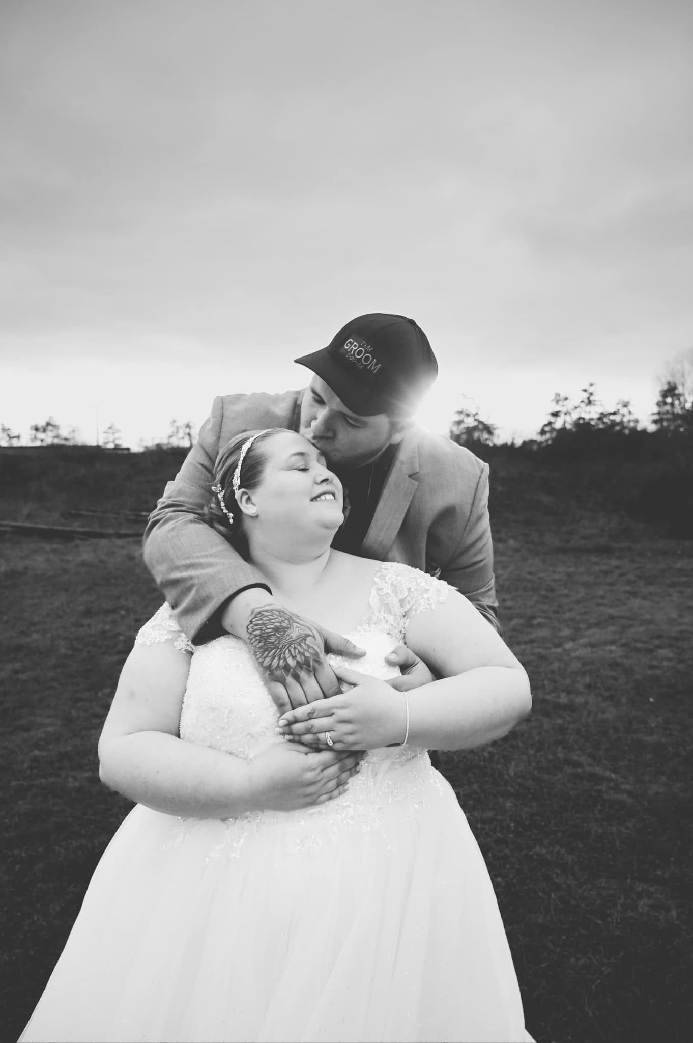 Black and white wedding photo of groom embracing bride from behind, kissing her cheek at sunset in an outdoor setting.