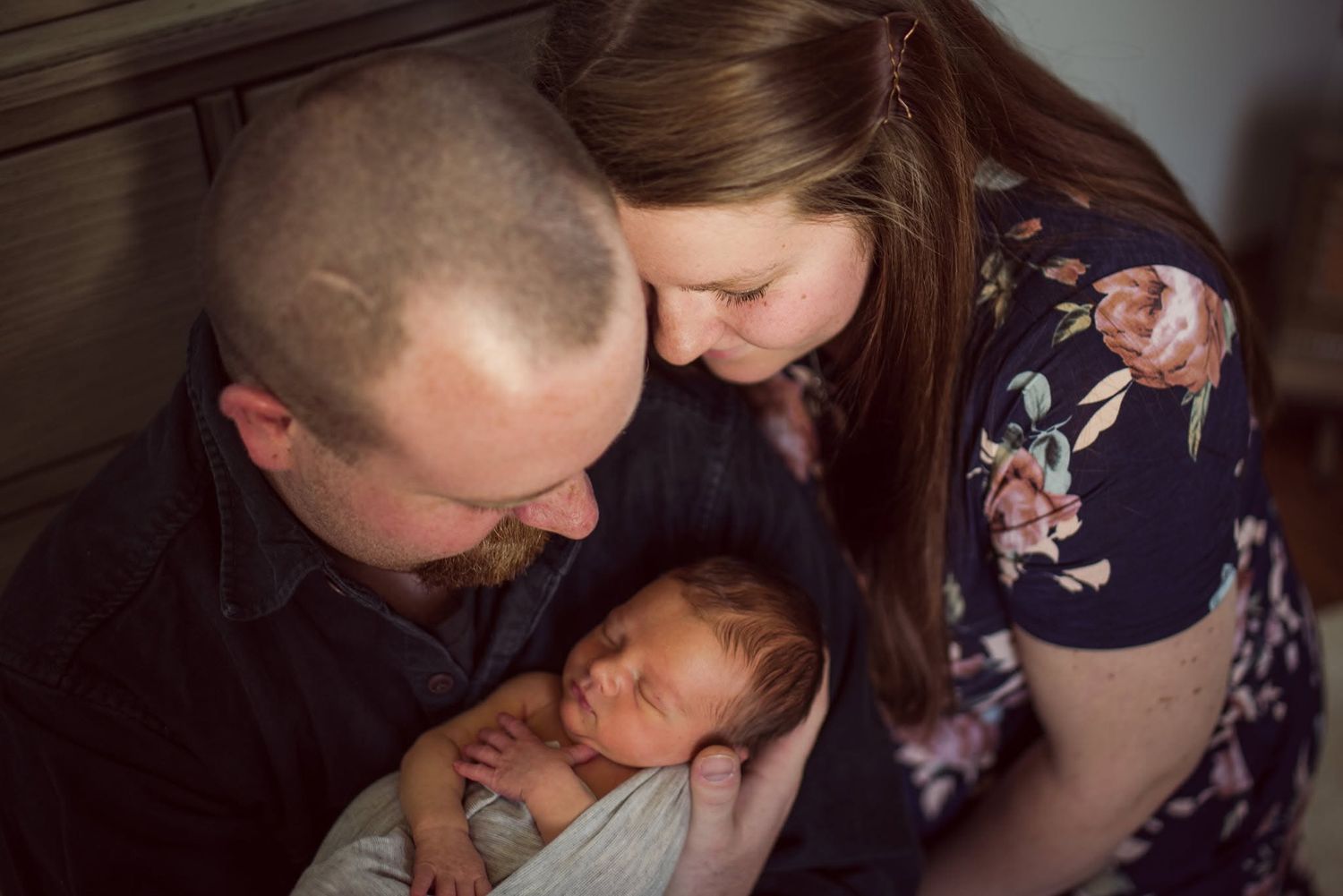 New parents lovingly gaze at their sleeping newborn baby wrapped in a white blanket during a newborn photo session.