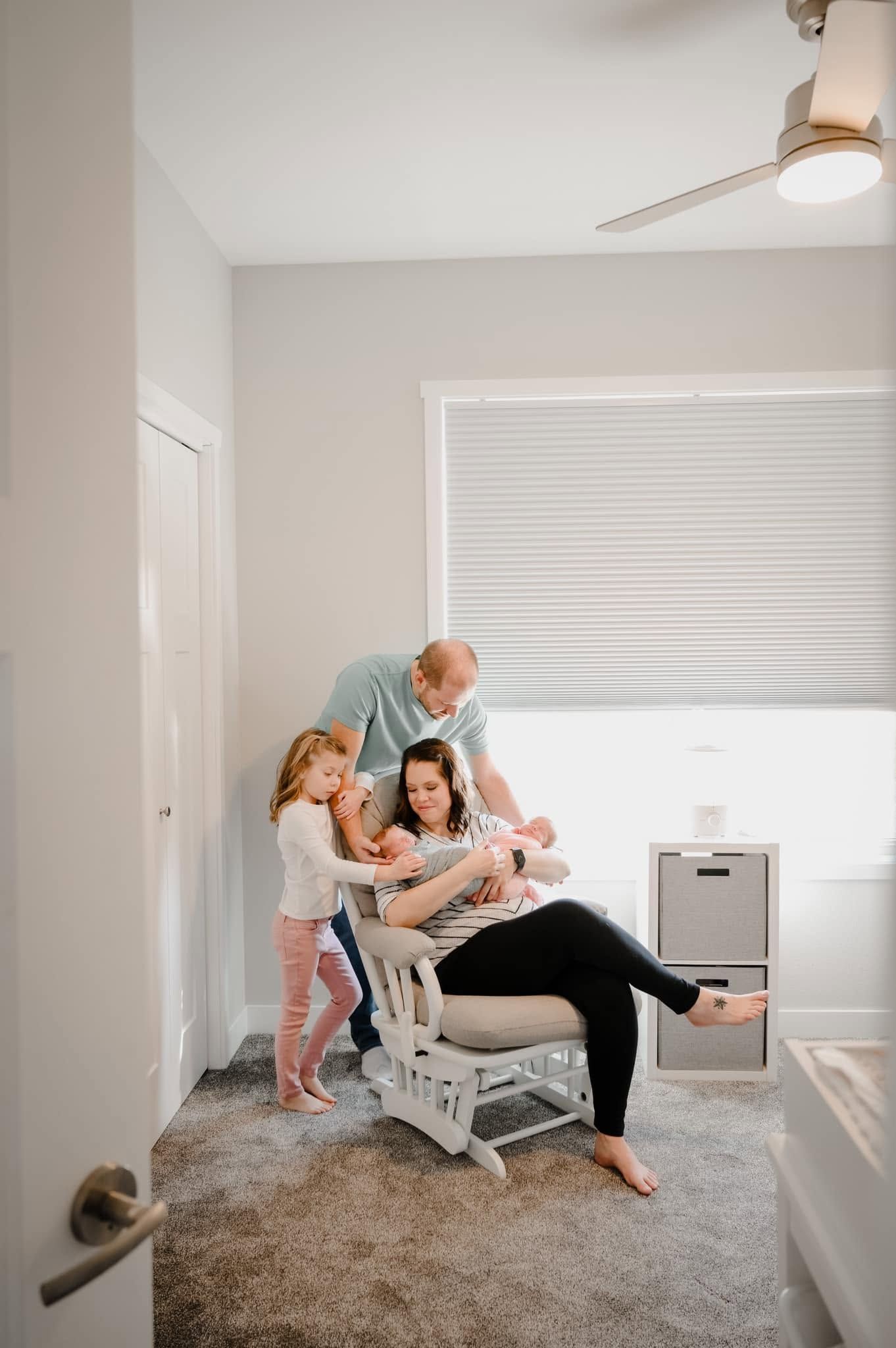 A family of four gathers in a nursery, with mom sitting in a glider holding a newborn while dad and toddler look on.