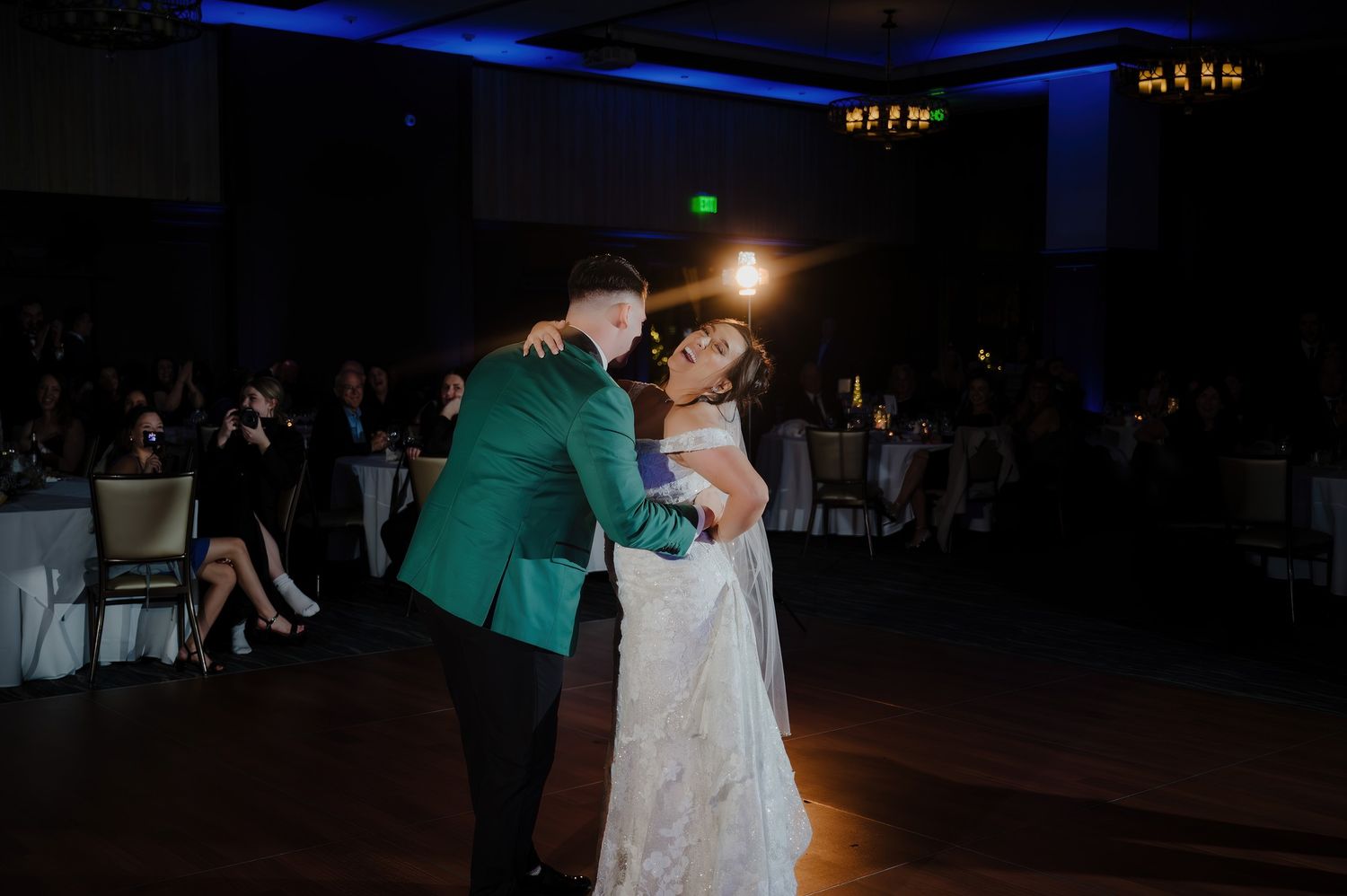 Couple sharing first dance at wedding reception, groom in green jacket, bride in white gown, blue ambient lighting.