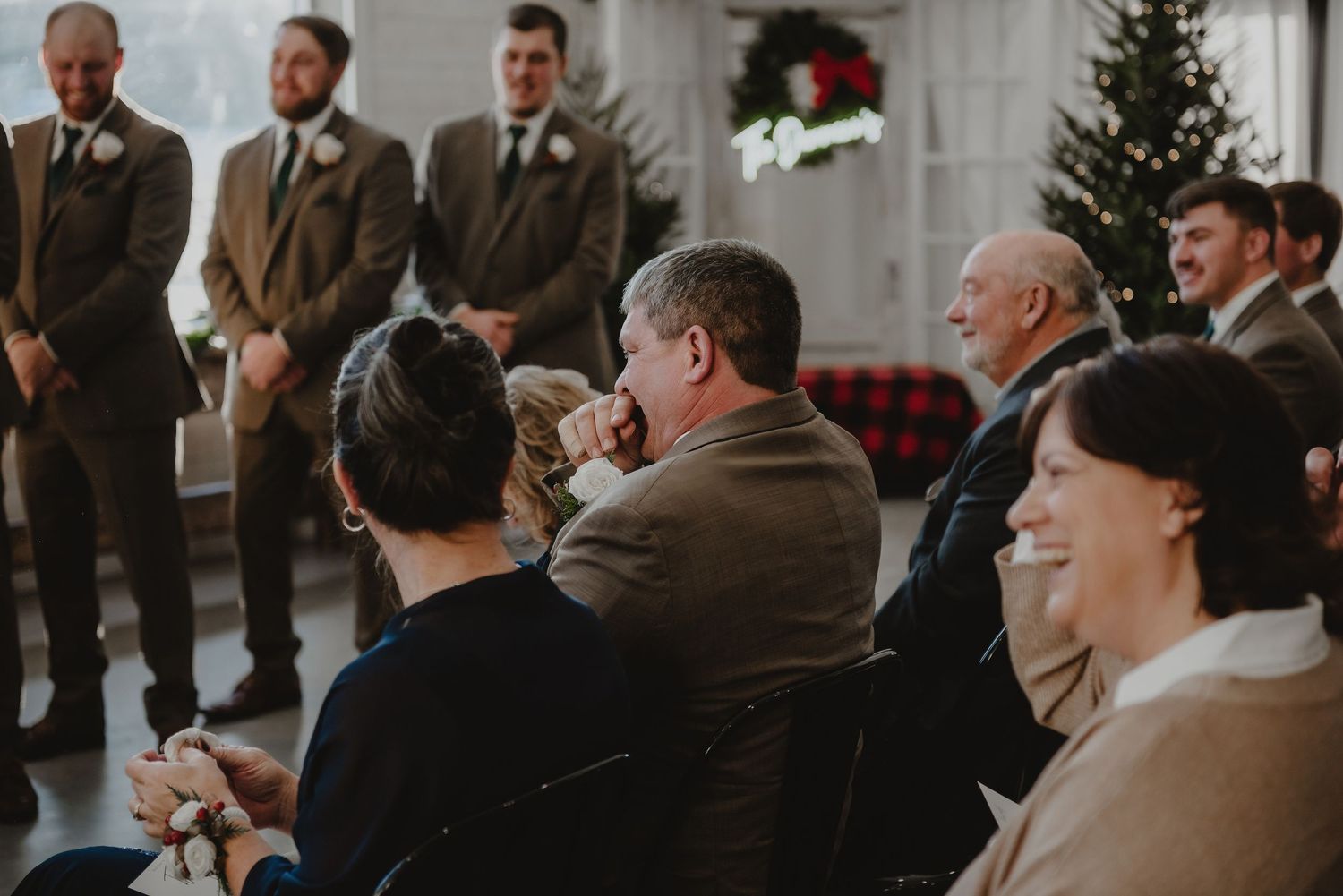 Wedding guests smile and laugh during an indoor ceremony with groomsmen standing near a decorated Christmas tree.