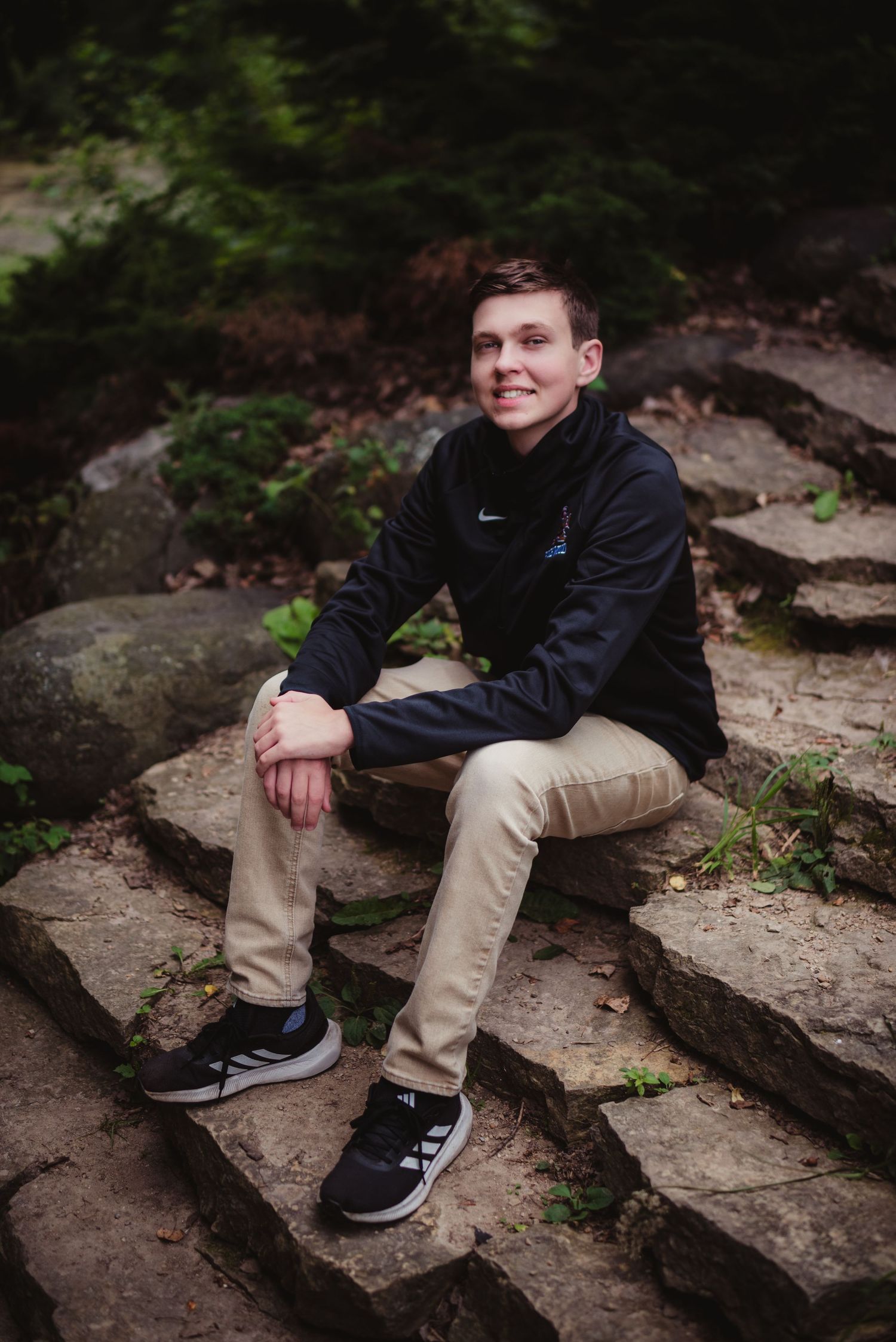 A smiling teenage boy sits on stone steps outdoors, wearing a Nike jacket, khaki pants, and Adidas sneakers.