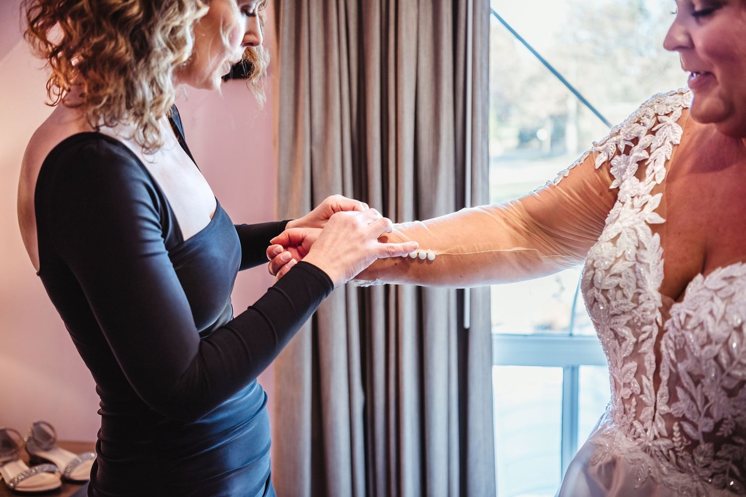Bridesmaid in navy dress helping bride in lace wedding gown with ring preparation indoors.