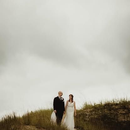 A bride and groom stand hand in hand on a grassy hill under a dramatic overcast sky on their wedding day.