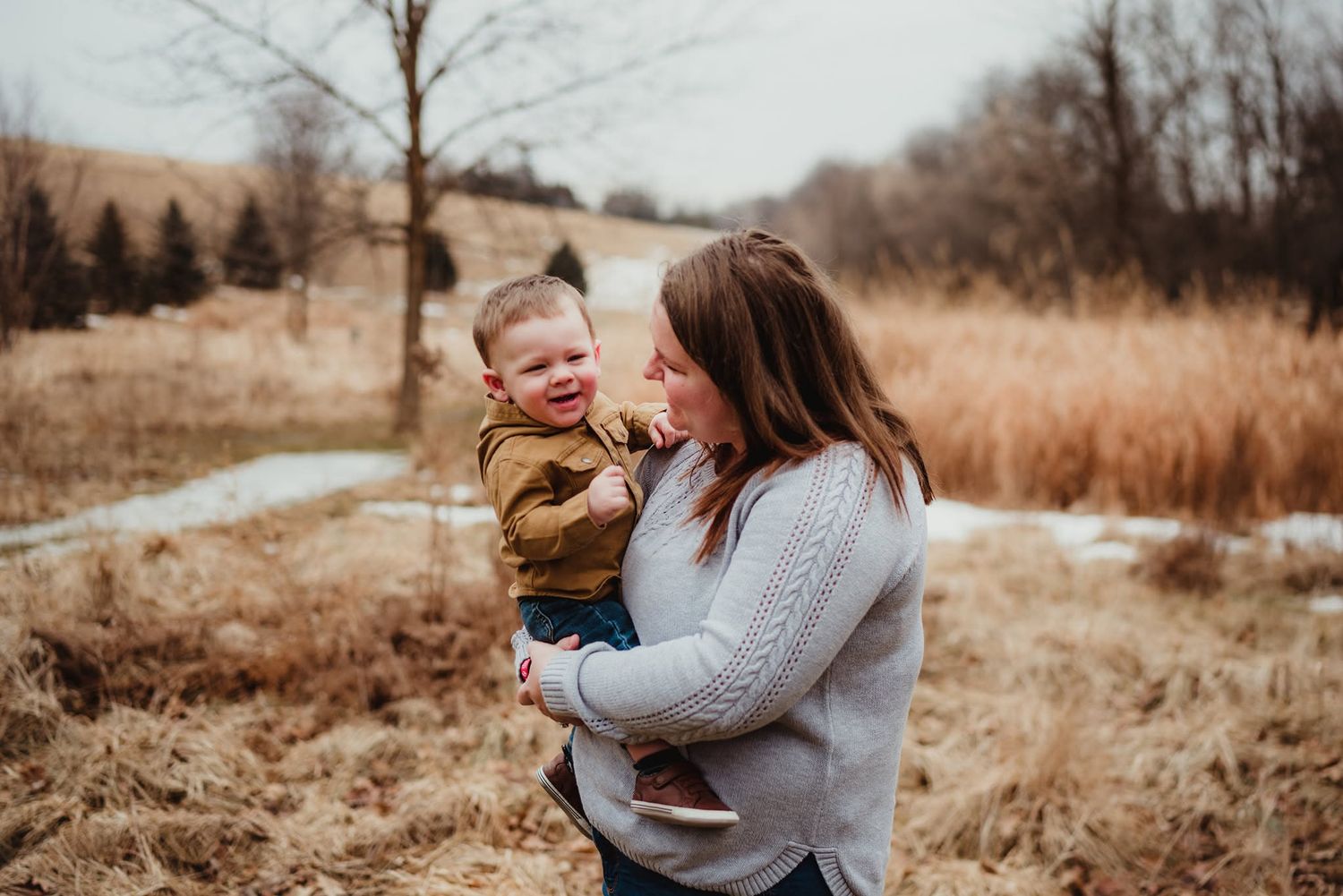 A mother holds her laughing toddler outdoors in a field with dry grass and bare trees in winter.