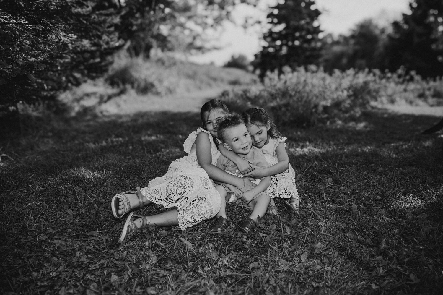 Black and white photo of three young children hugging and playing together on grass outdoors.