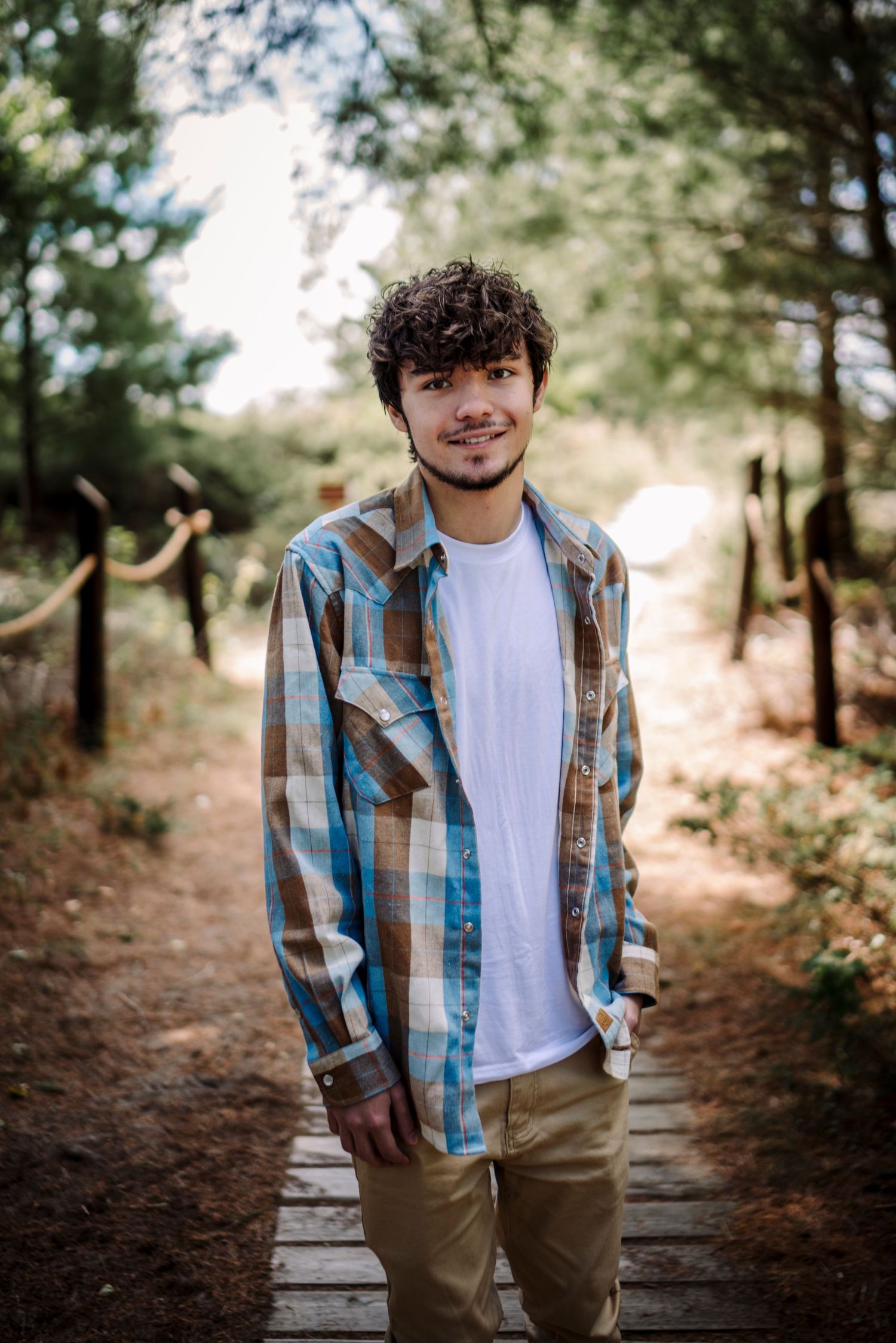 Young man with curly hair smiling on a wooden path in a forest, wearing a plaid shirt over a white tee.