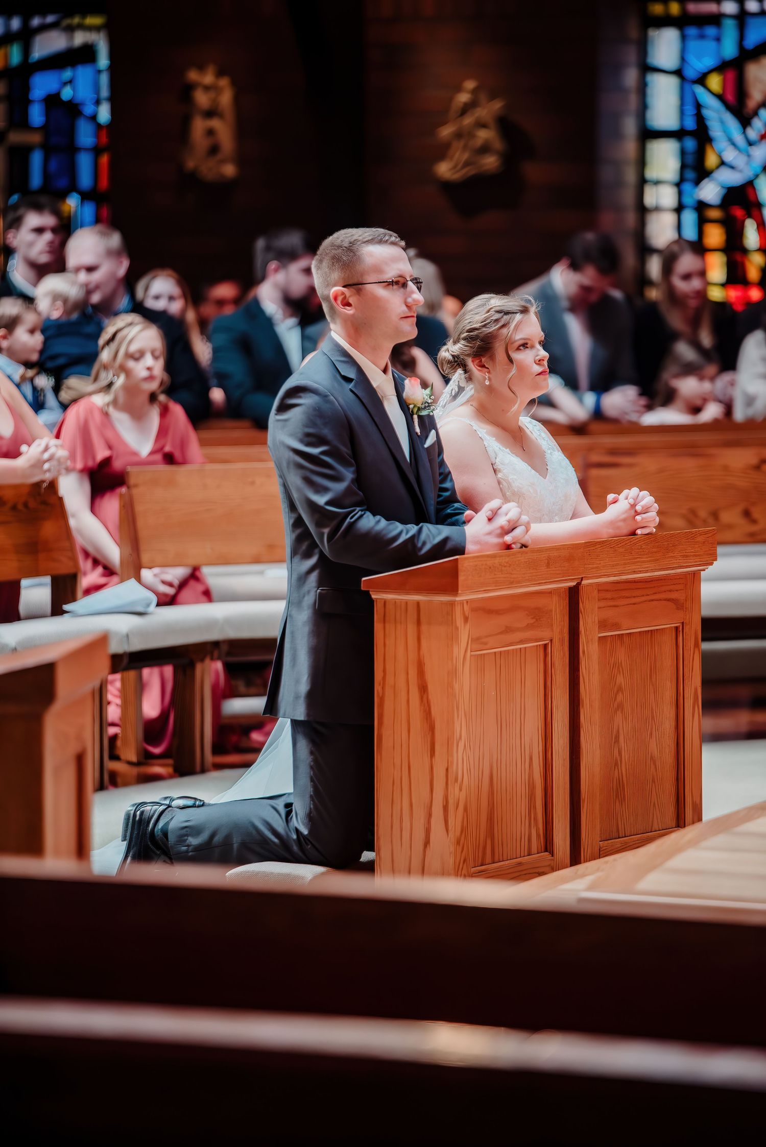 Bride and groom kneeling in prayer at wooden prie-dieu during Catholic wedding ceremony in church.