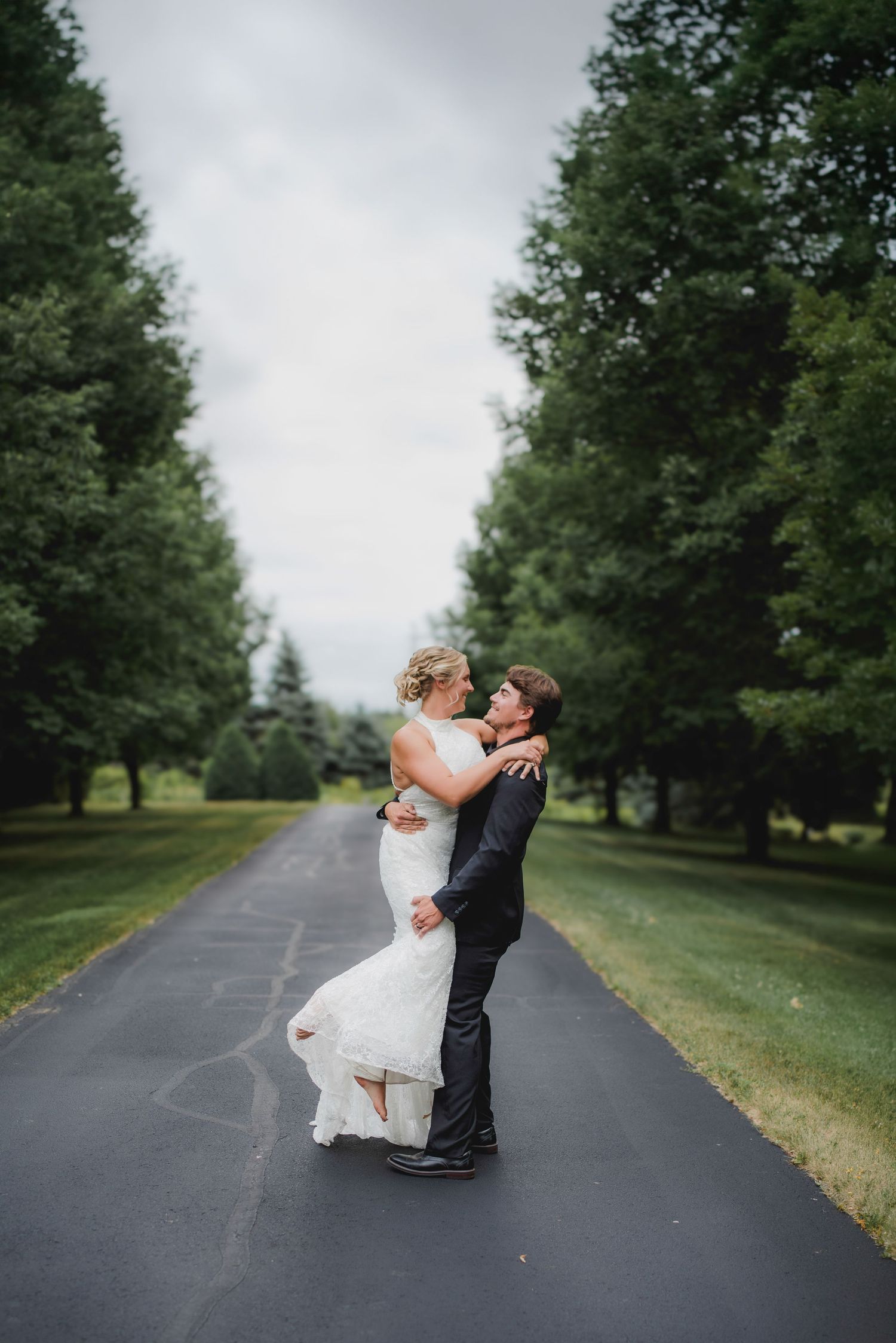 Bride and groom embracing on a tree-lined driveway in a romantic outdoor wedding portrait.