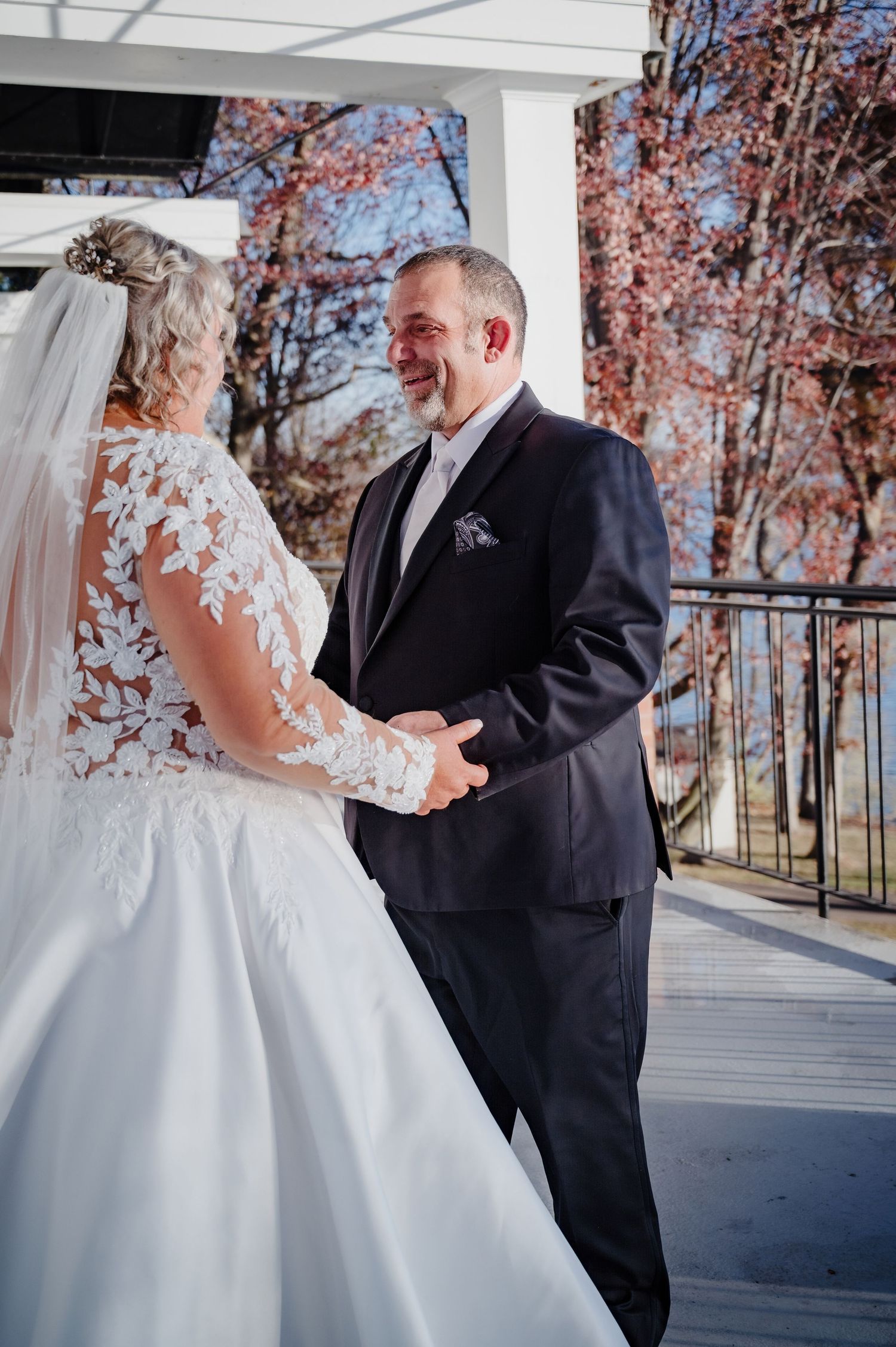 A groom in a dark suit smiles joyfully at his bride in a lace wedding gown on a decorated outdoor venue.