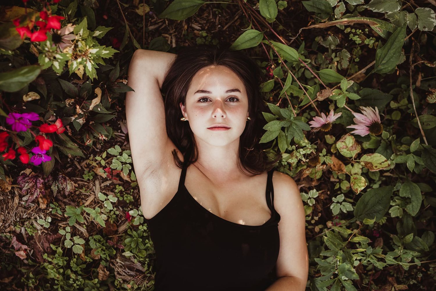 Young woman lying among colorful garden flowers and green leaves, looking up serenely in natural light.