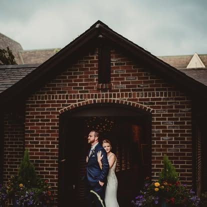 A bride and groom pose romantically in front of a rustic brick chapel entrance adorned with colorful flowers.