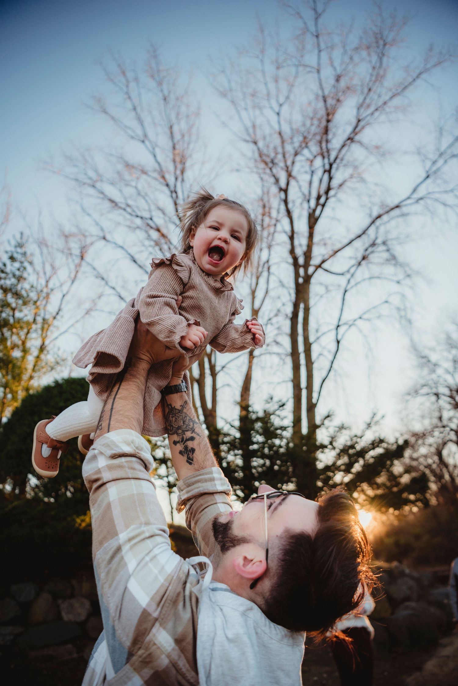 Parent joyfully lifting laughing toddler in the air outdoors at sunset with bare trees in background.