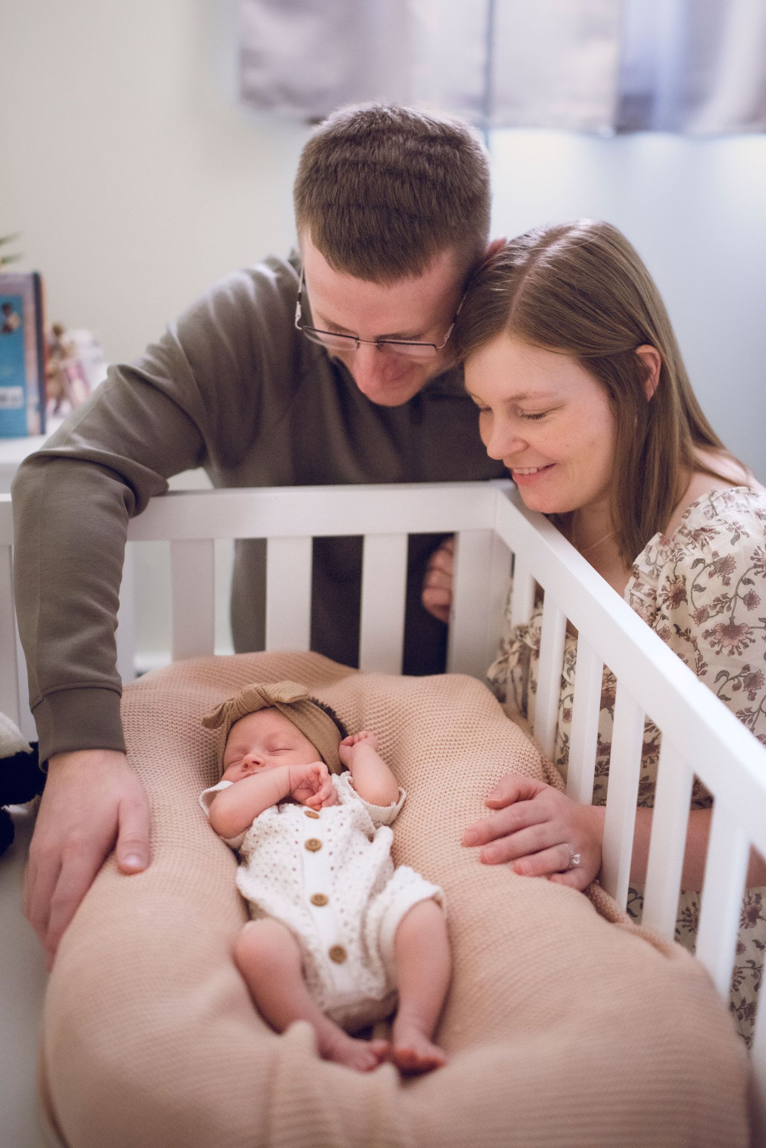 New parents lovingly gaze at their sleeping newborn baby in a white crib, sharing a tender family moment.