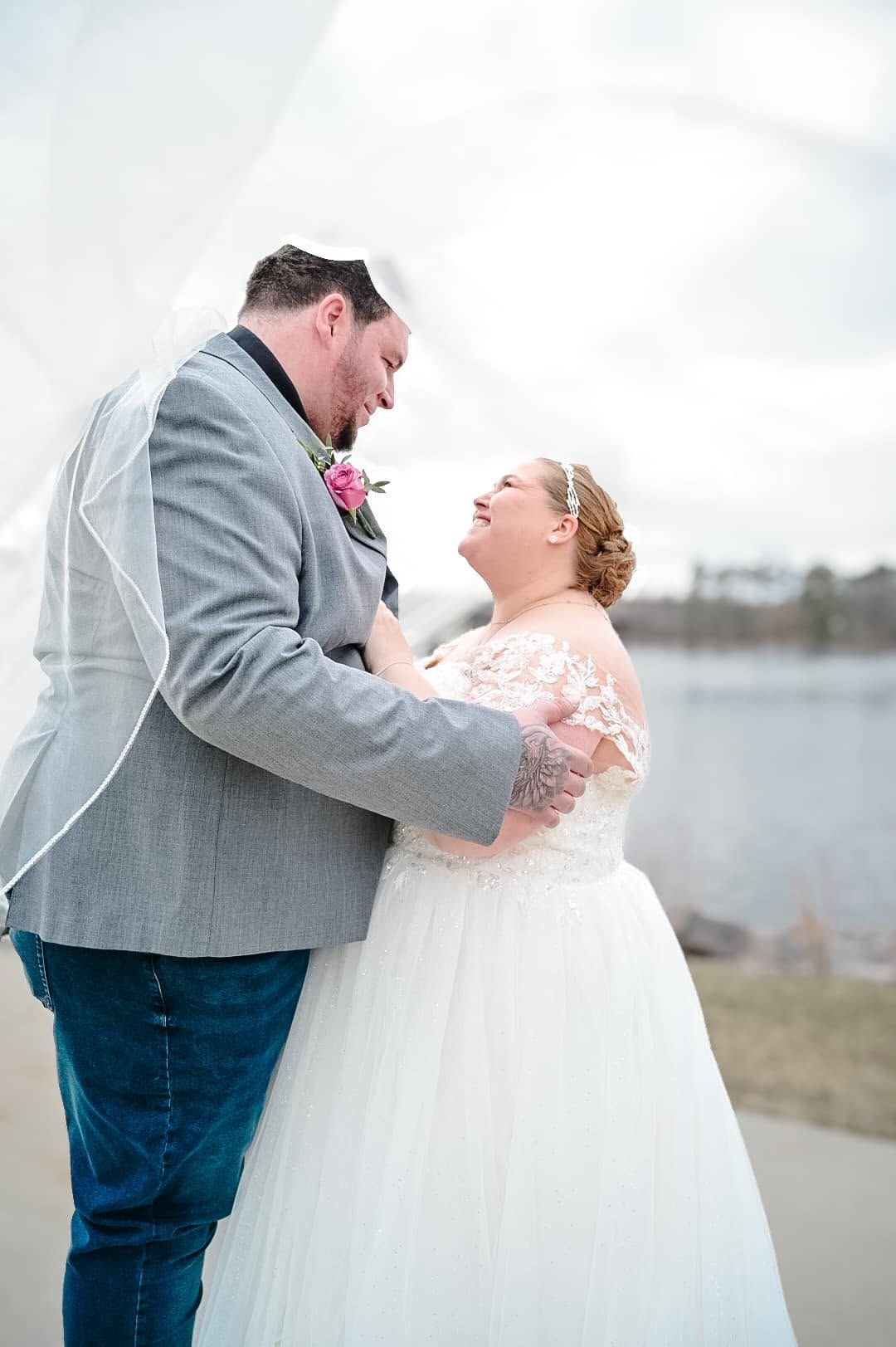 A joyful bride and groom share a romantic moment outdoors near a lake, with the bride laughing in a lace wedding gown.