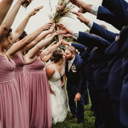 Wedding party tunnel with bridesmaids in dusty rose gowns and groomsmen in navy suits as bride and groom share a kiss.