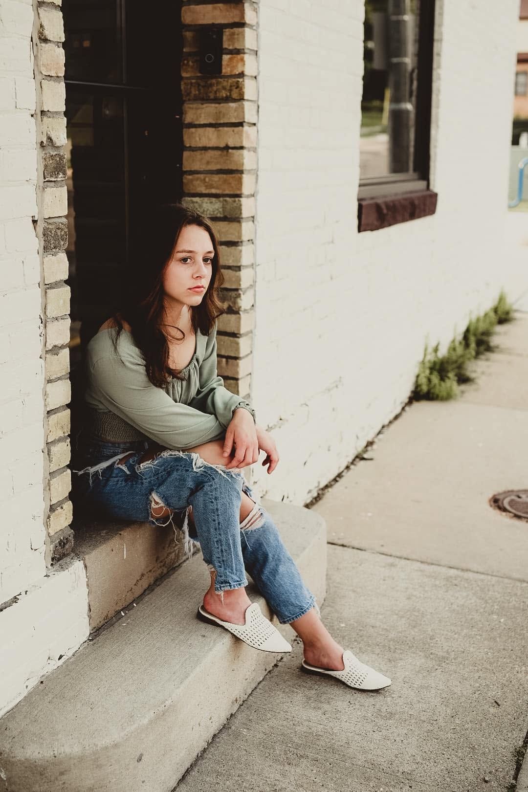 Young woman in ripped jeans and green top sits pensively on building steps beside brick wall.