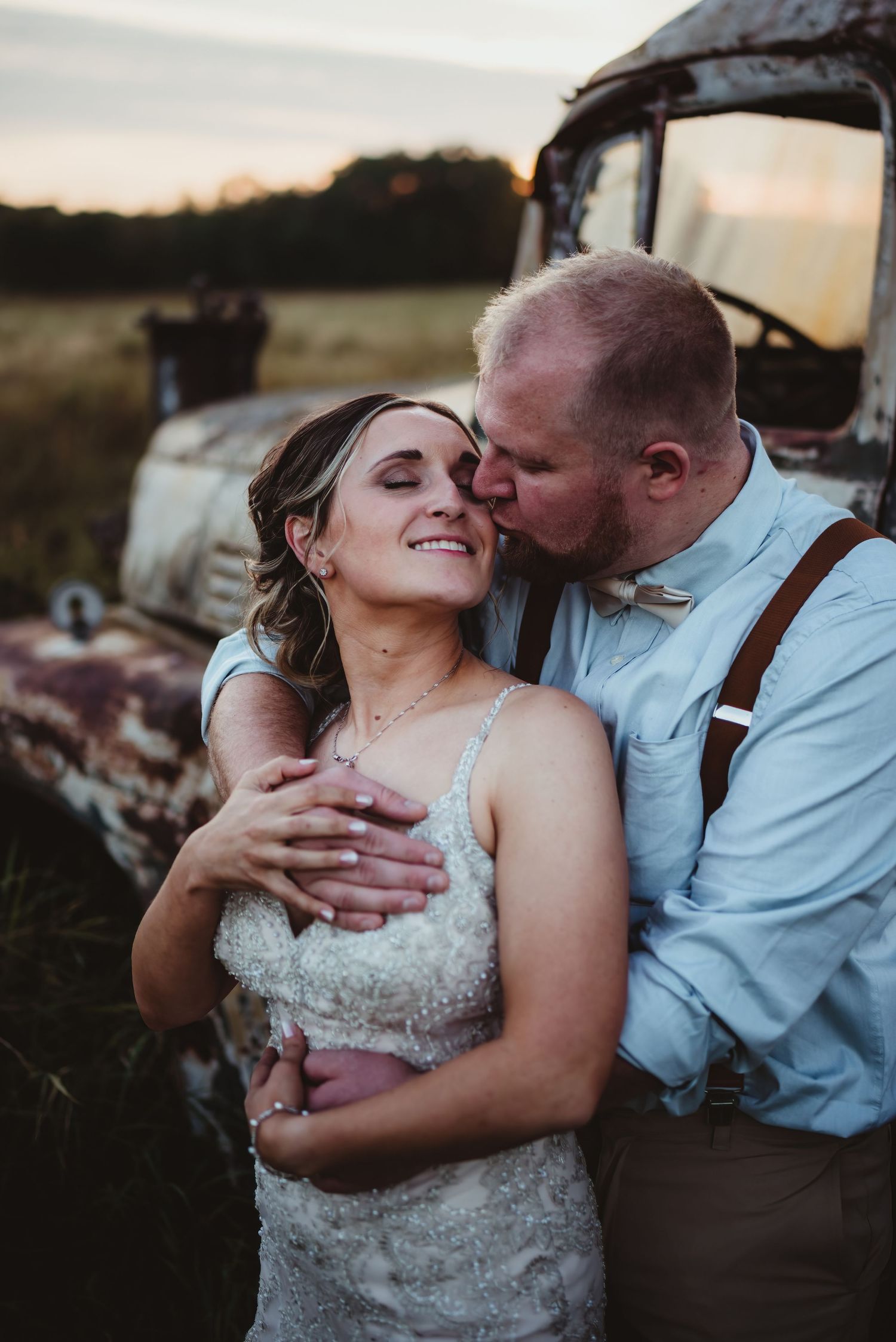 Groom in suspenders kissing bride in lace wedding dress near rustic truck at sunset.