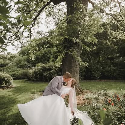A bride and groom share a romantic dip kiss in a lush garden setting beneath a large tree.