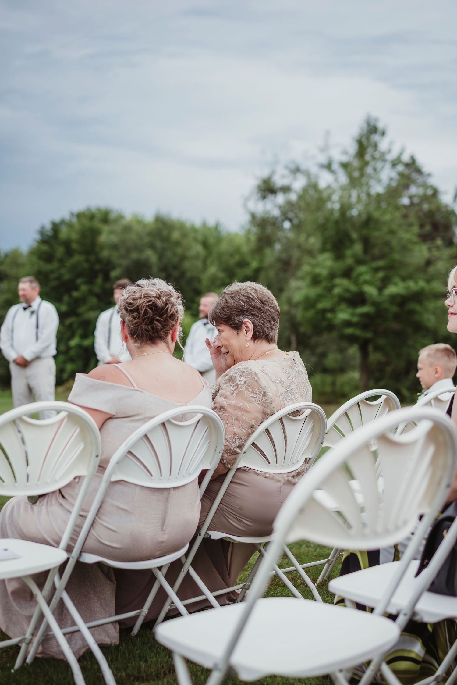 Guests seated in white folding chairs at an outdoor wedding ceremony, surrounded by lush green trees.