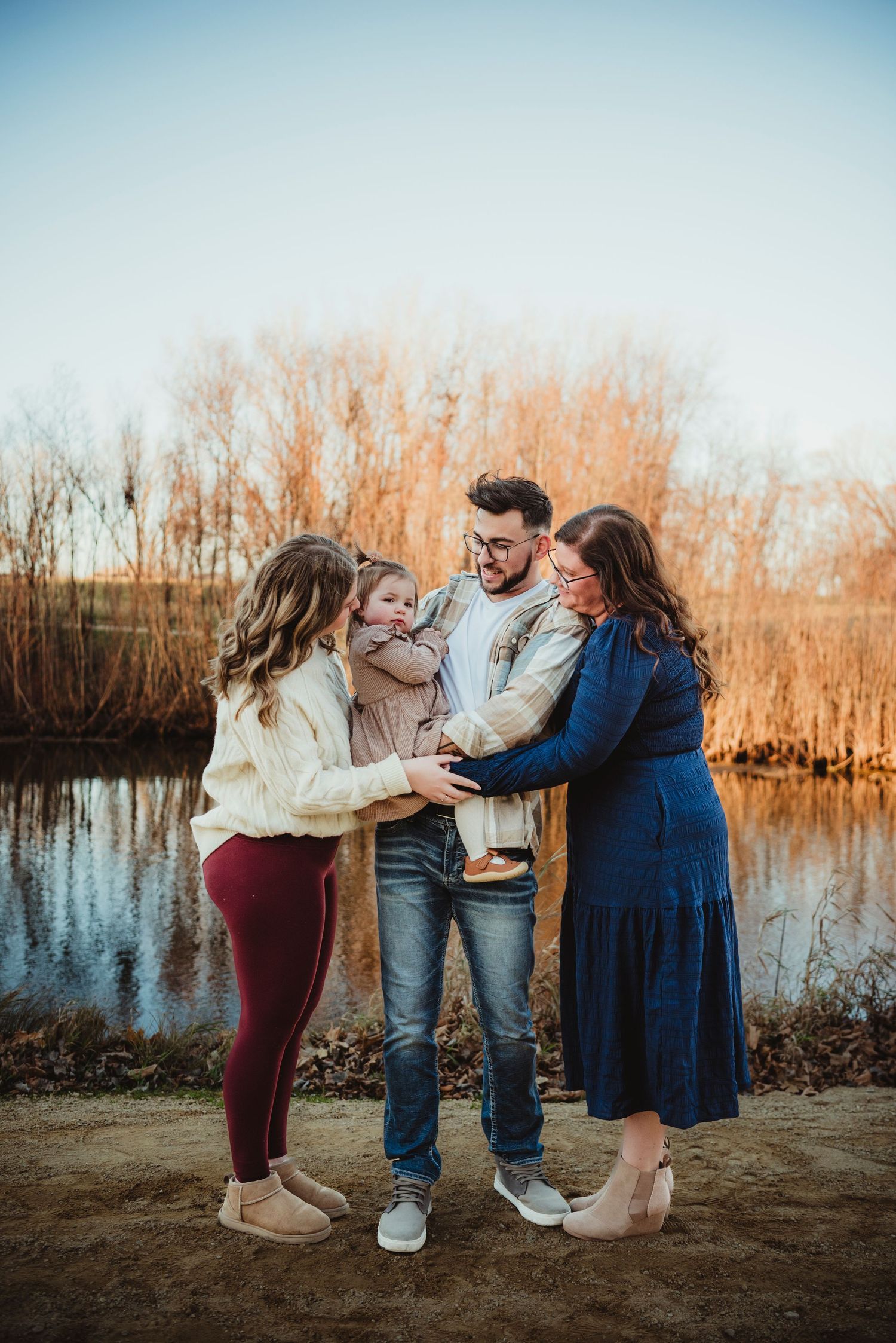 Family of three sharing a tender moment by a reflective pond at golden hour, parents holding young child.
